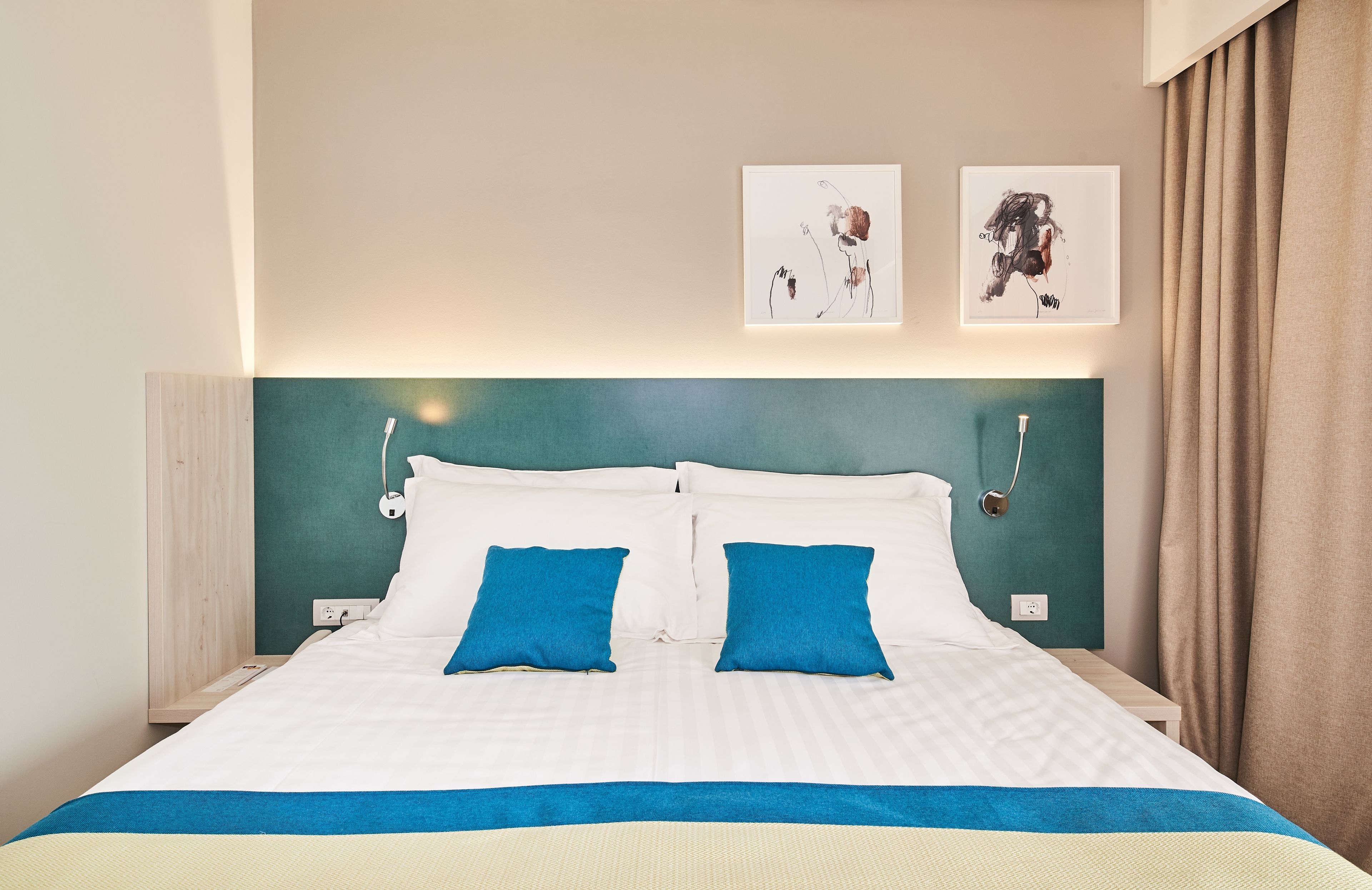 Una cama bien hecha con ropa de cama blanca y azul, dos obras de arte colgadas en la pared y un cabecero color turquesa.