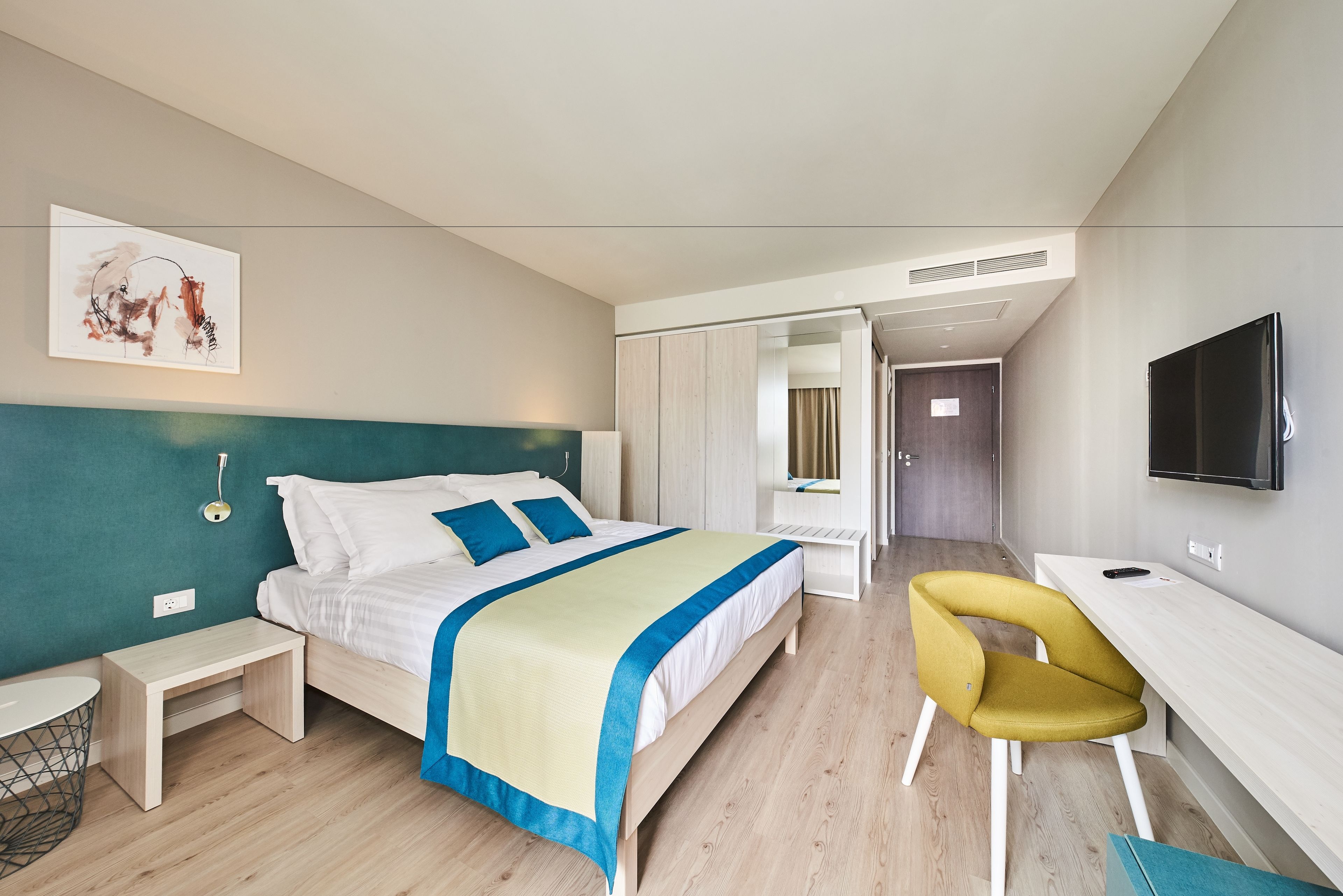 A modern hotel room with a bed, bedside table, chair, desk, and TV.