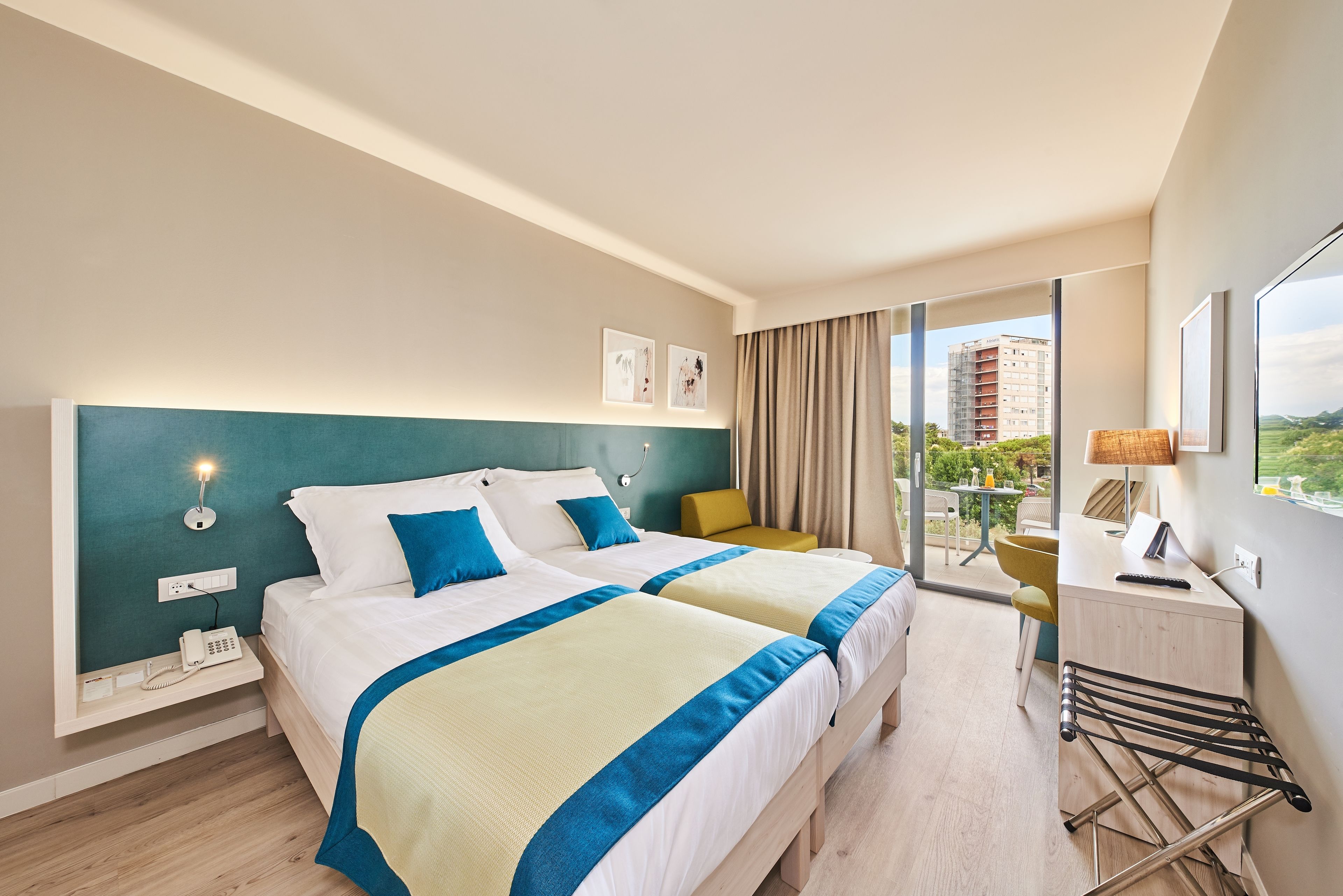 Une chambre d’hôtel avec deux lits, un bureau, une chaise et un balcon donnant sur des immeubles.