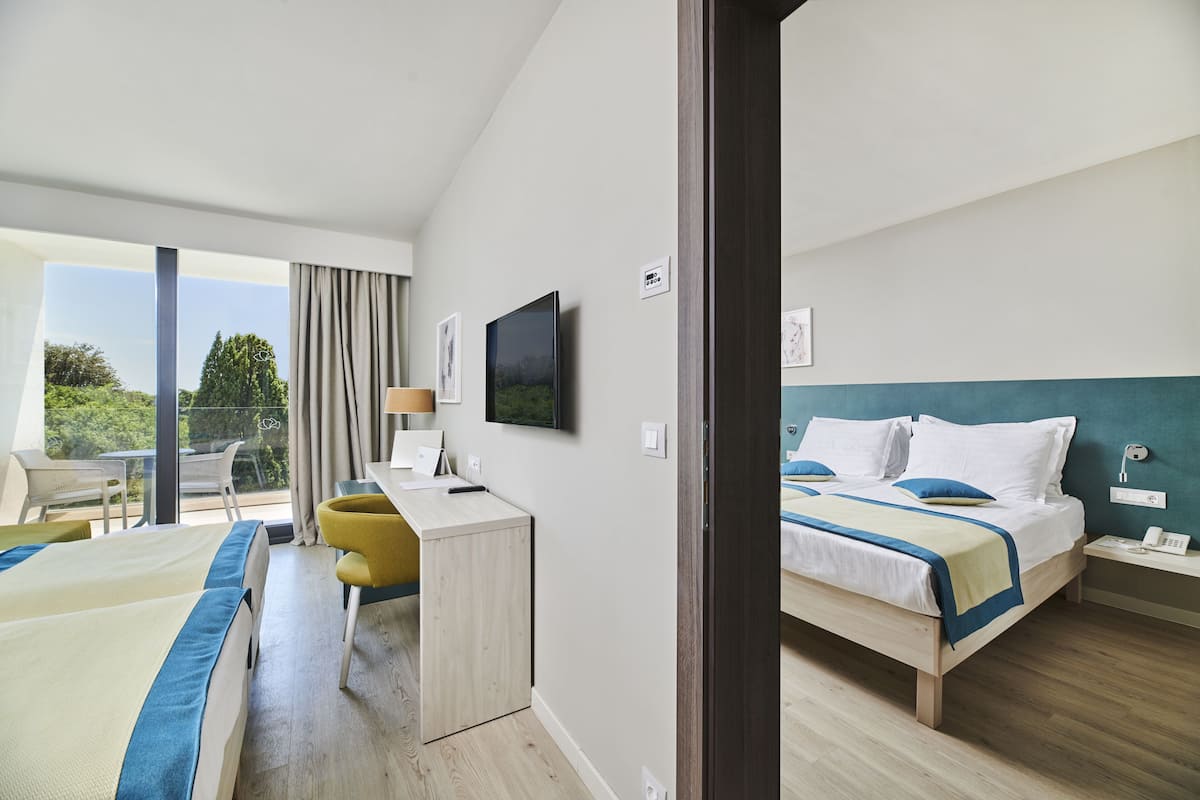 A hotel room with two beds, a desk, a chair, a TV, and a balcony with outdoor seating.