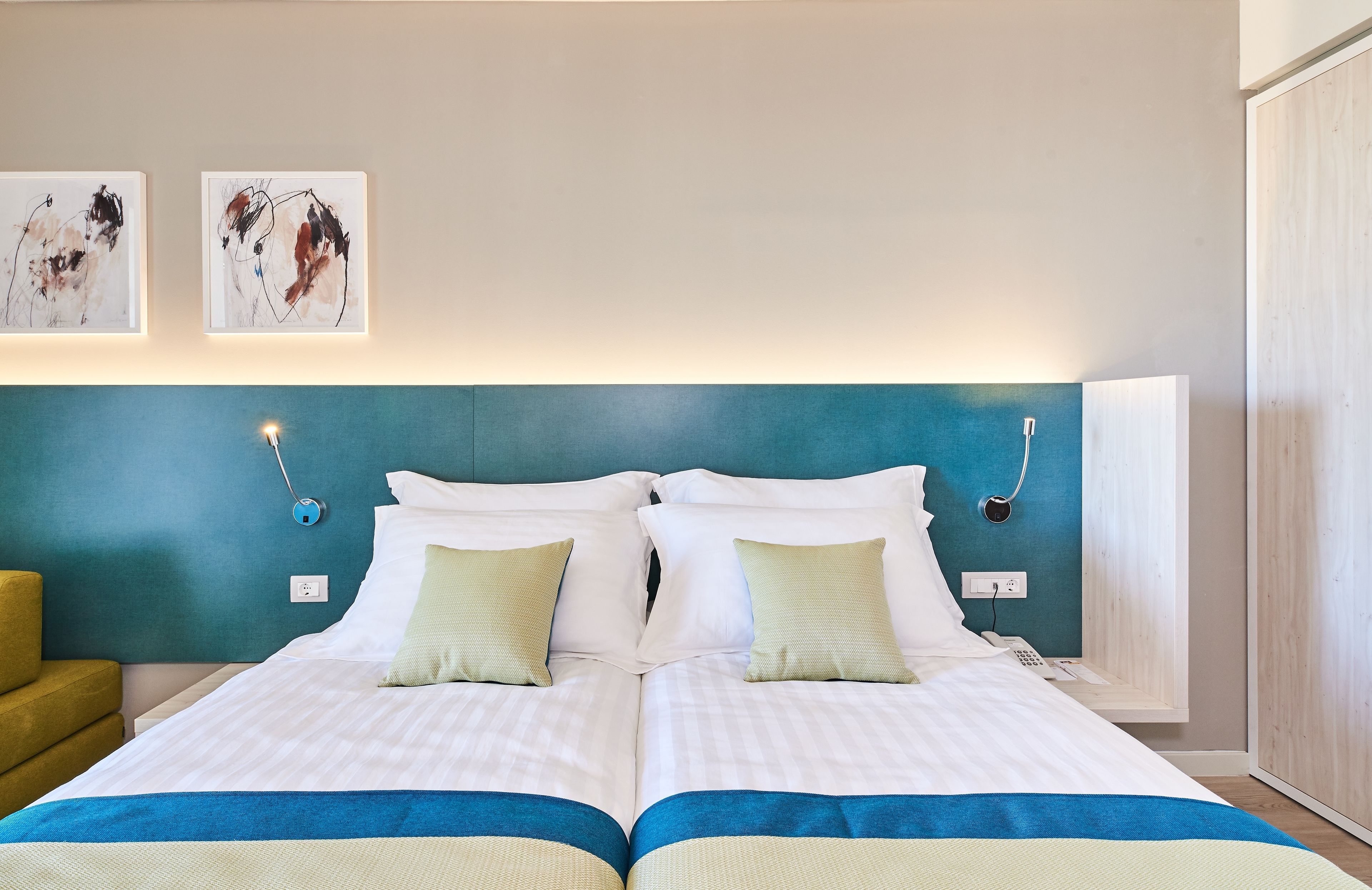 Una cama doble con cabecera color turquesa, ropa de cama blanca y dos almohadas verdes. En la pared sobre la cama hay pinturas abstractas.