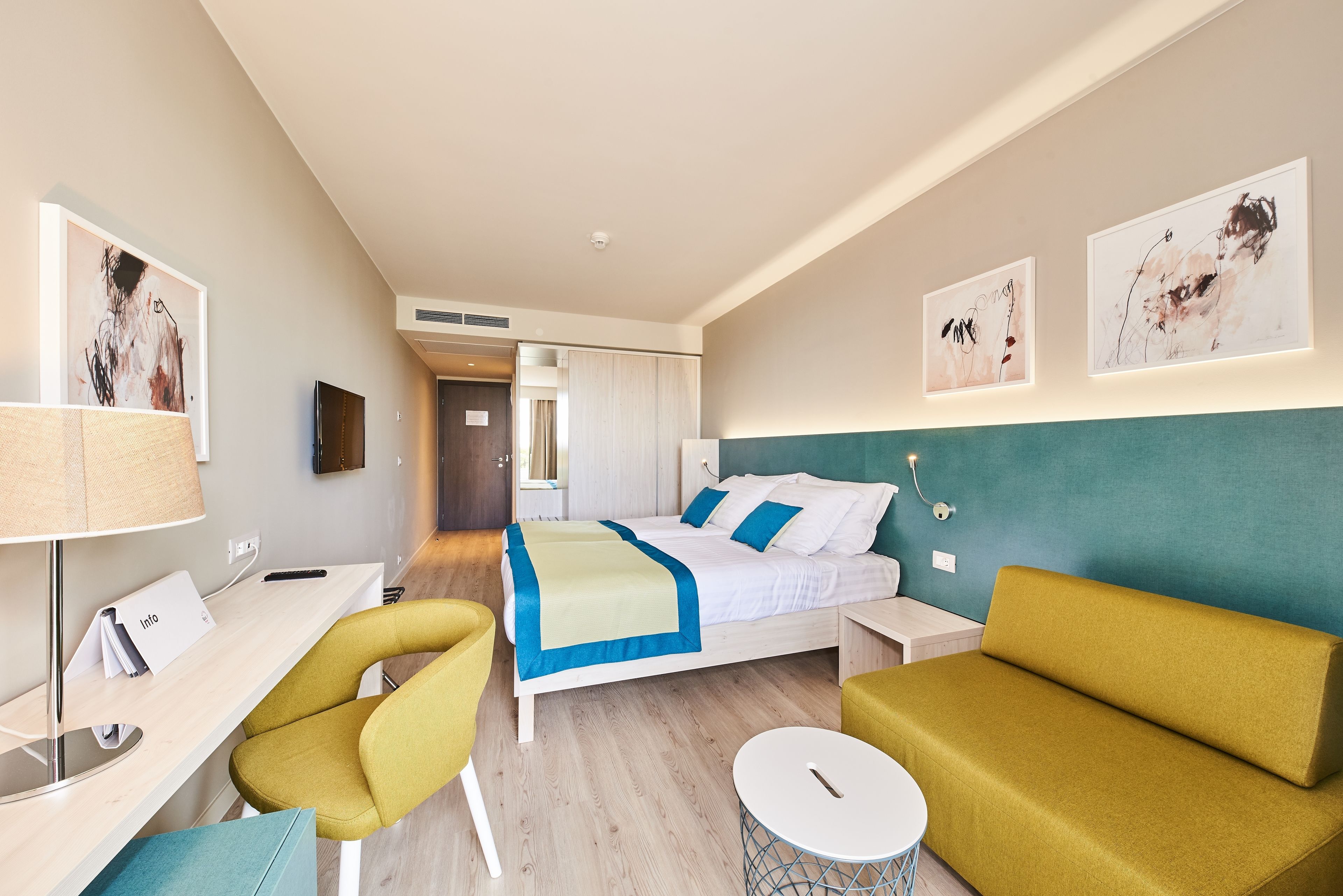 A hotel room with a bed, a desk, a chair, a sofa, and artwork on the walls.