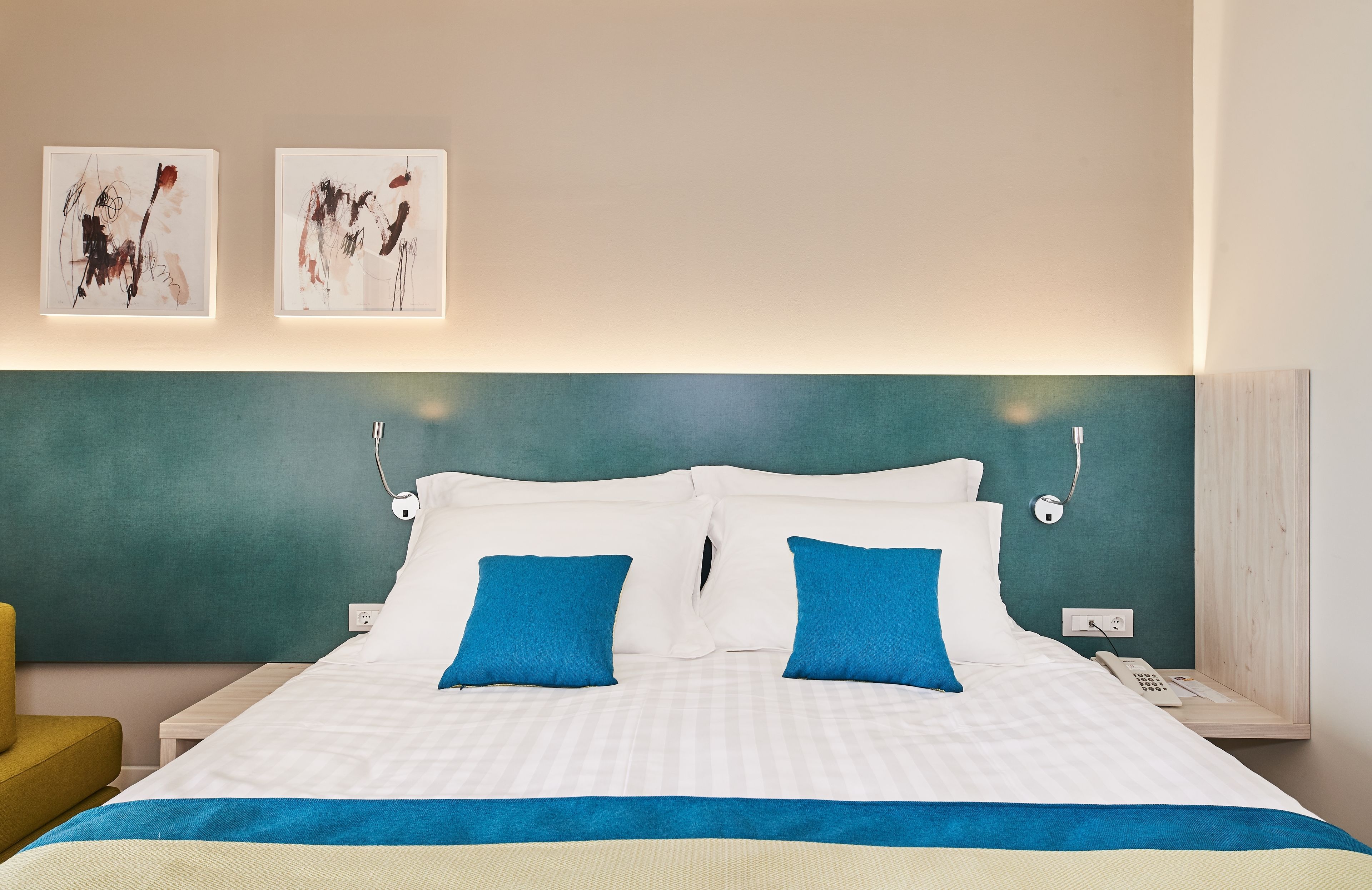 Een hotelkamer met een bed, twee blauwe kussens, een witte deken en abstracte kunst aan de muur.