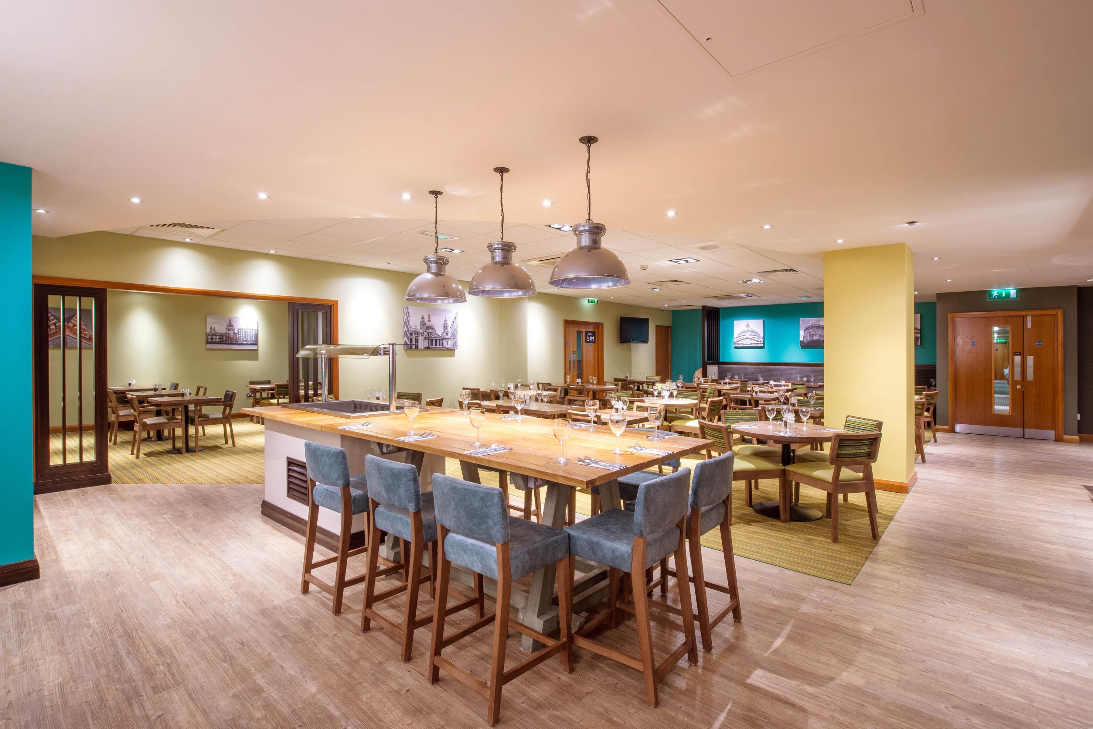 Uma área de refeições ampla, com várias mesas e cadeiras, iluminação pendente e um bar central.