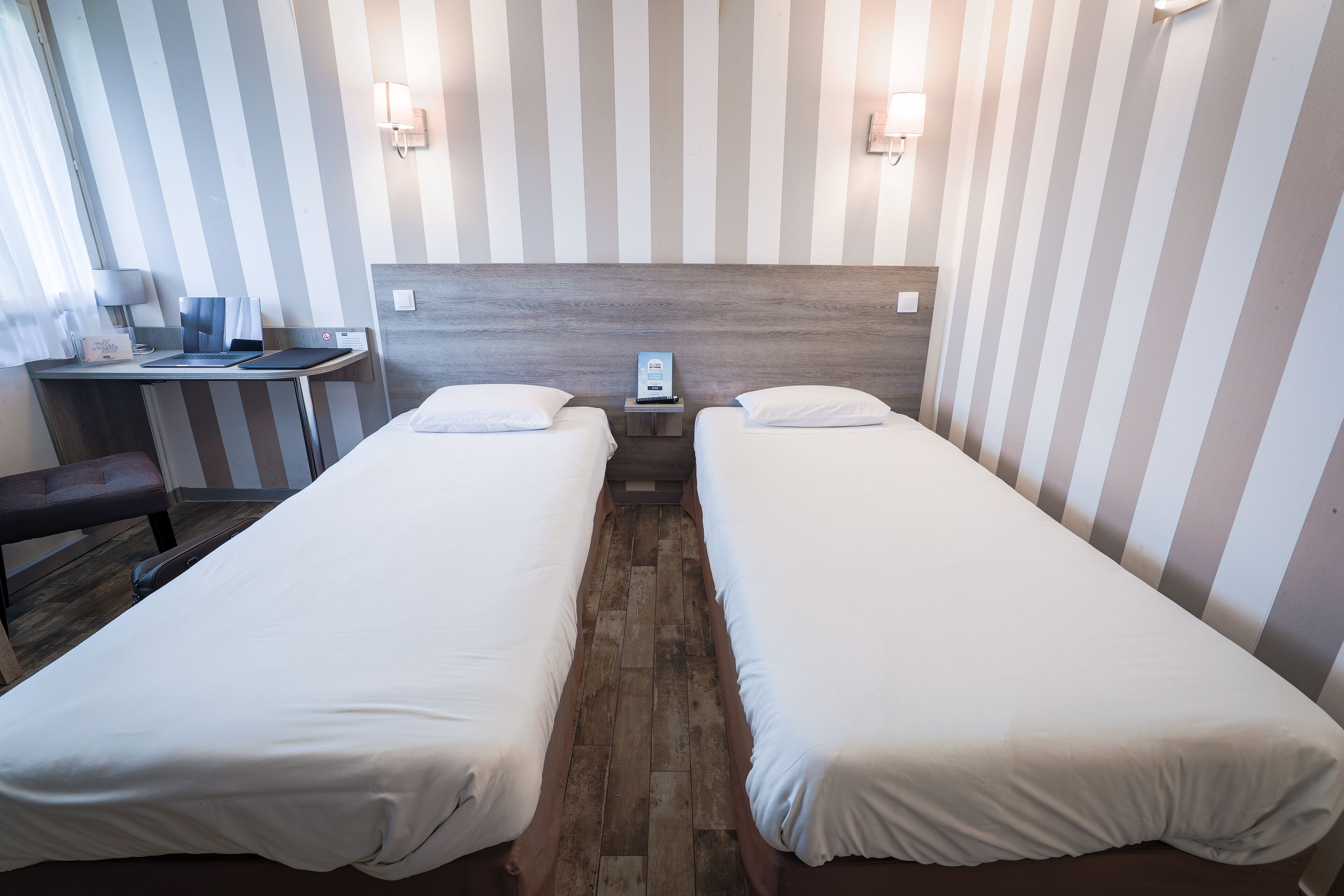 Una camera d'albergo con due letti singoli, una scrivania con un portatile, una sedia e un comodino con il telefono.