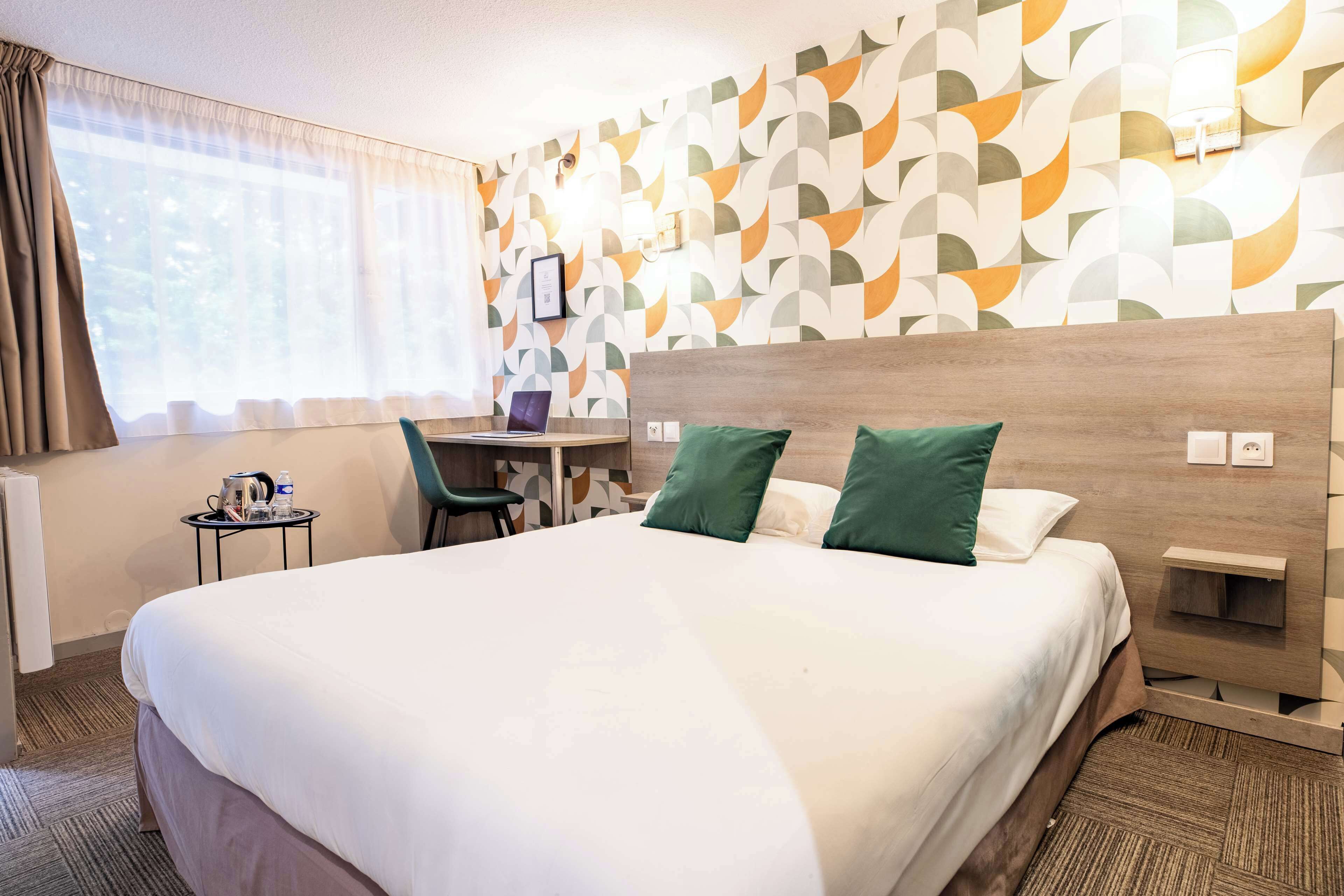 Habitación de hotel con una cama grande, un escritorio y una silla. Se ve una ventana con cortinas, una pared con un diseño geométrico y una lámpara de pared.