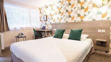 Habitación de hotel con una cama grande, un escritorio y una silla. Se ve una ventana con cortinas, una pared con un diseño geométrico y una lámpara de pared.