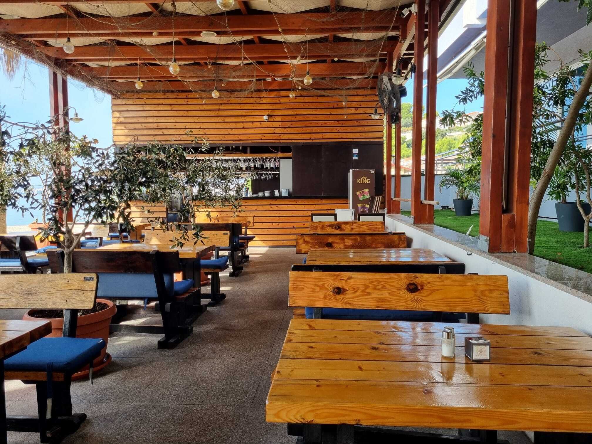 Un café al aire libre con mesas y bancos de madera, plantas en macetas y vista al mar.