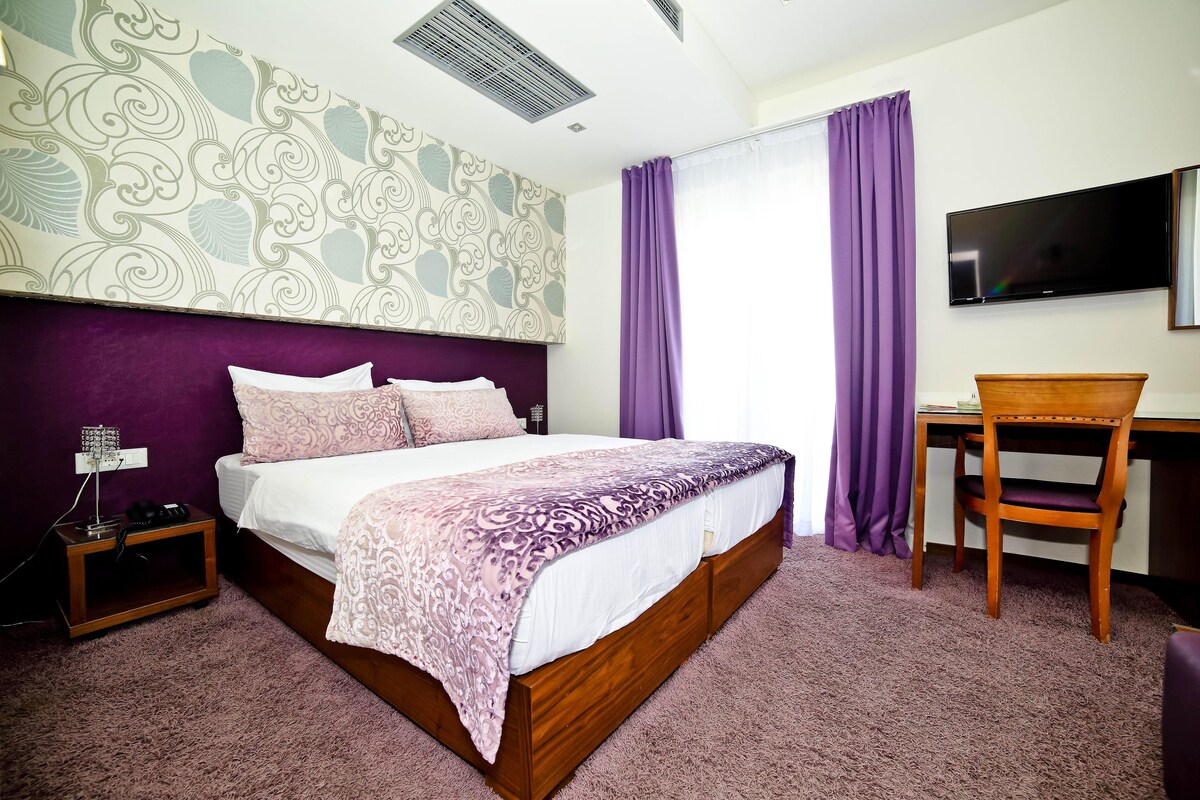 A bedroom with a bed, a desk, a chair, a TV, and a patterned wall.