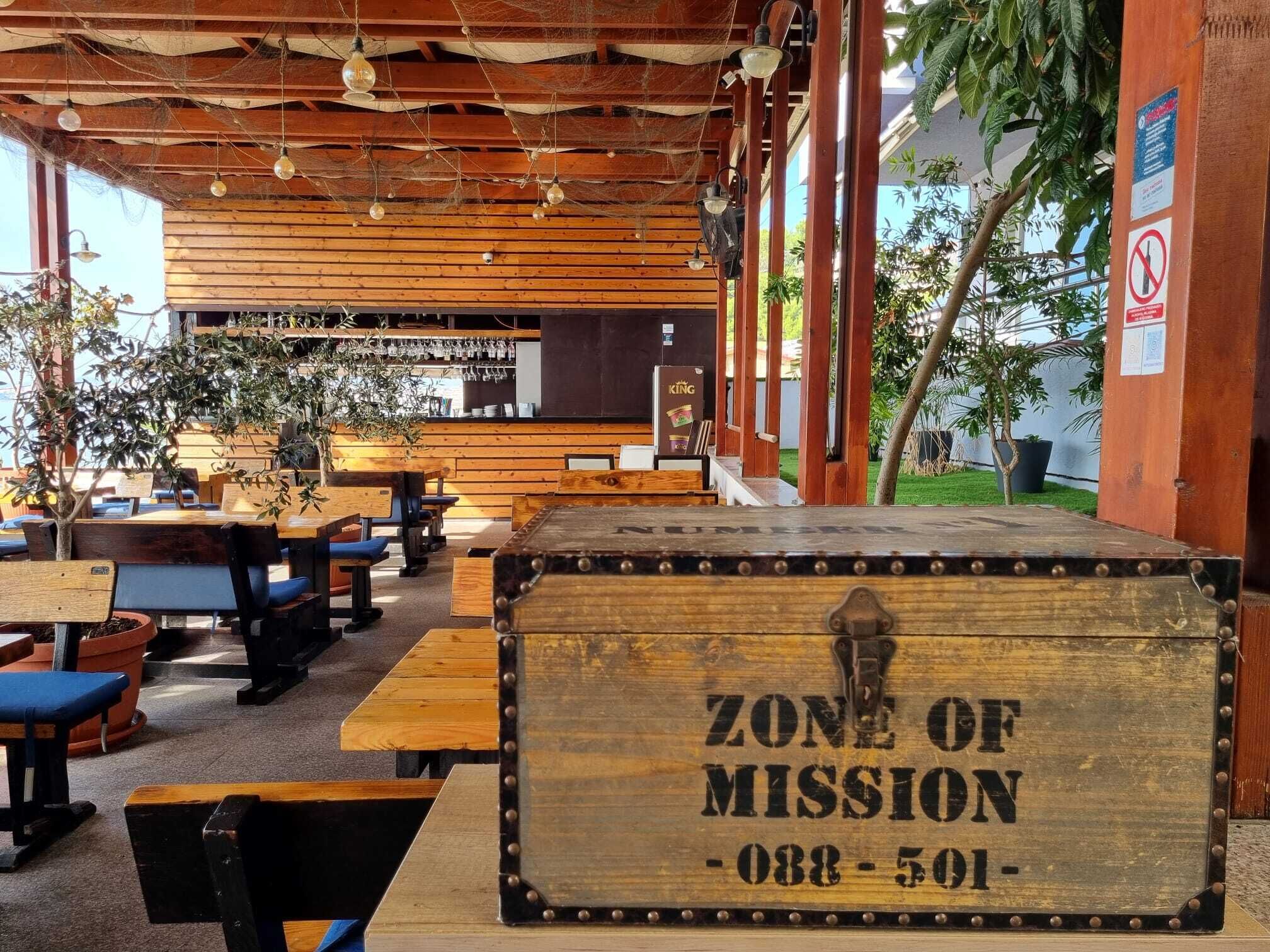 Et rustikt udendørs opholdsområde med træborde og bænke, et træskilt med teksten ”ZONE OF MISSION - 088-501-”, og et barområde i baggrunden.