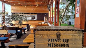 Et rustikt udendørs opholdsområde med træborde og bænke, et træskilt med teksten ”ZONE OF MISSION - 088-501-”, og et barområde i baggrunden.
