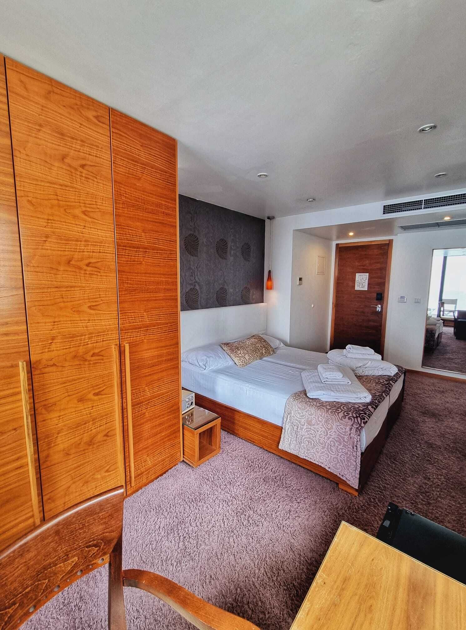 A hotel room with a wooden wardrobe, a bed with a patterned bedspread, a small bedside table, and a mirror.