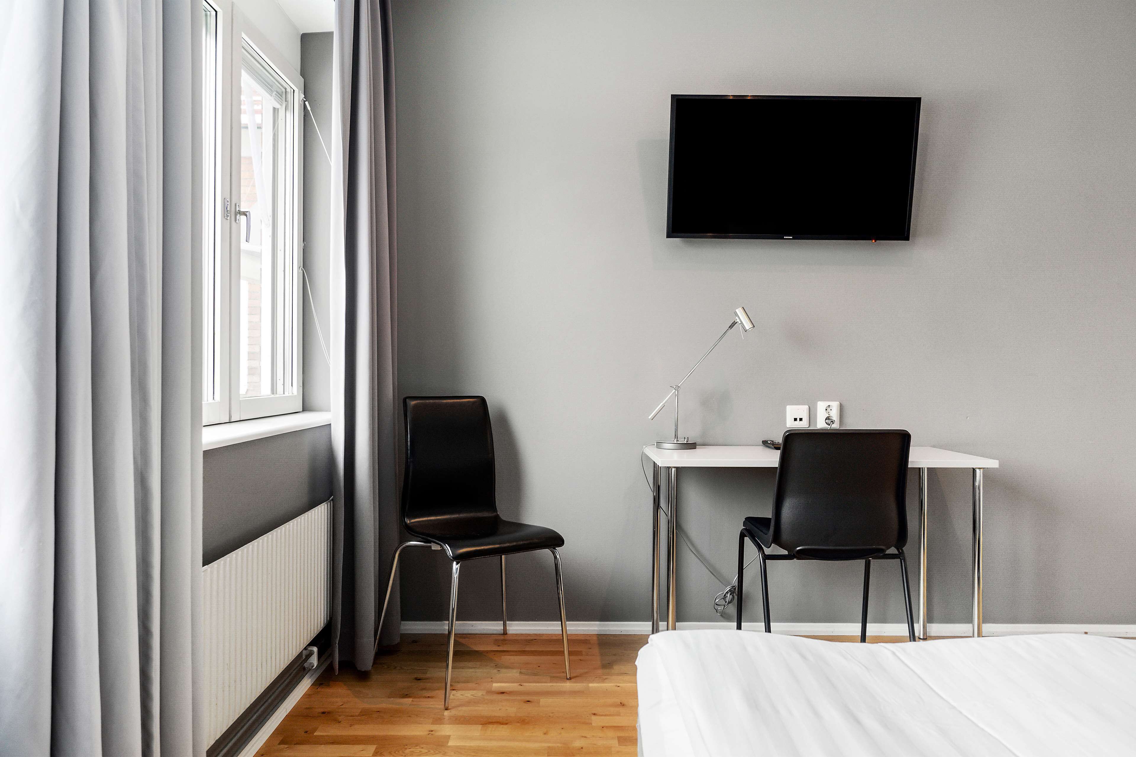 Ett hotellrum med en säng, ett skrivbord med två stolar, en tv som är monterad på väggen och ett fönster med gardiner.