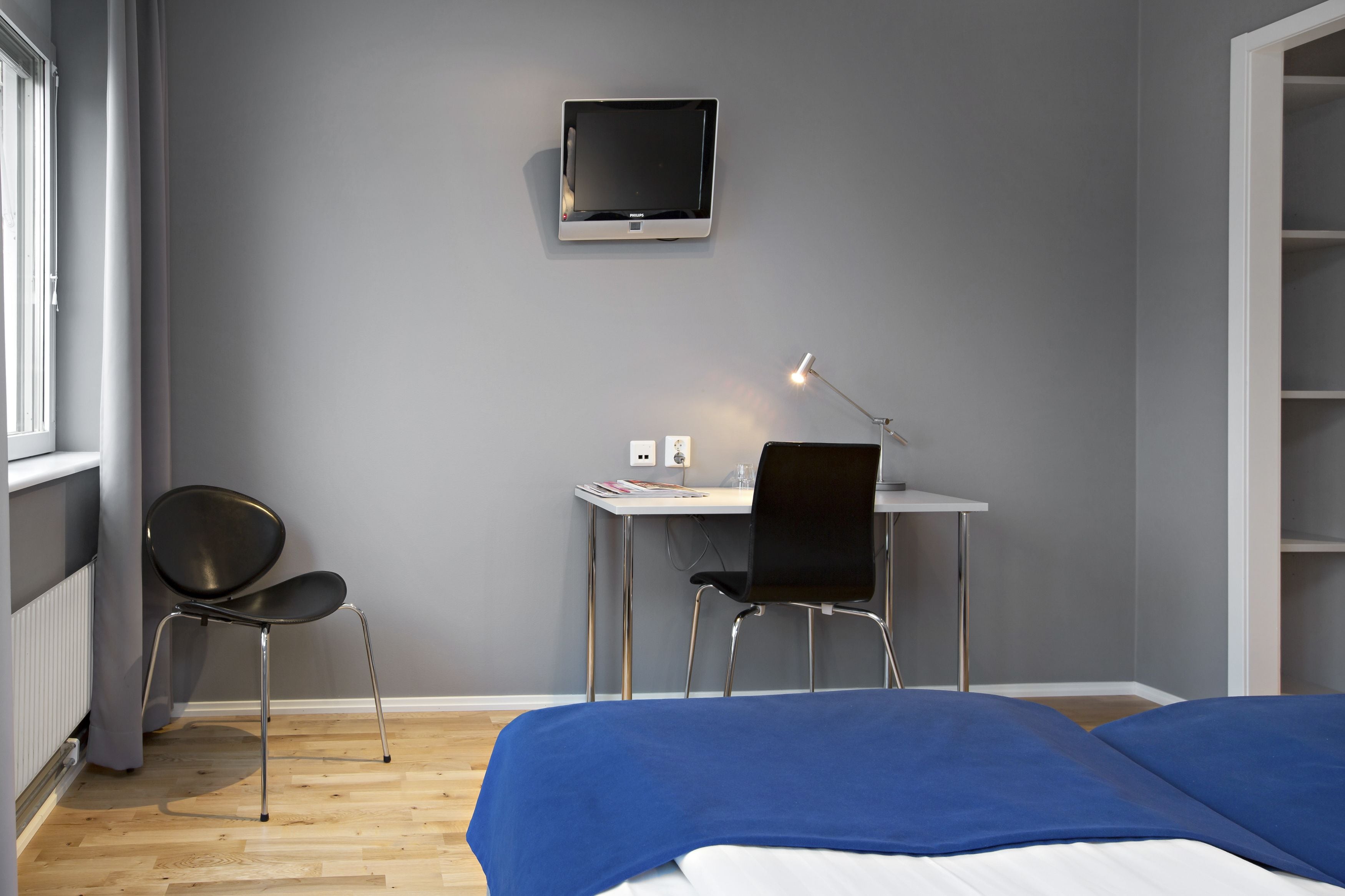 Een slaapkamer met een bed, een bureau met stoel, een aan de muur bevestigde televisie en een lamp.