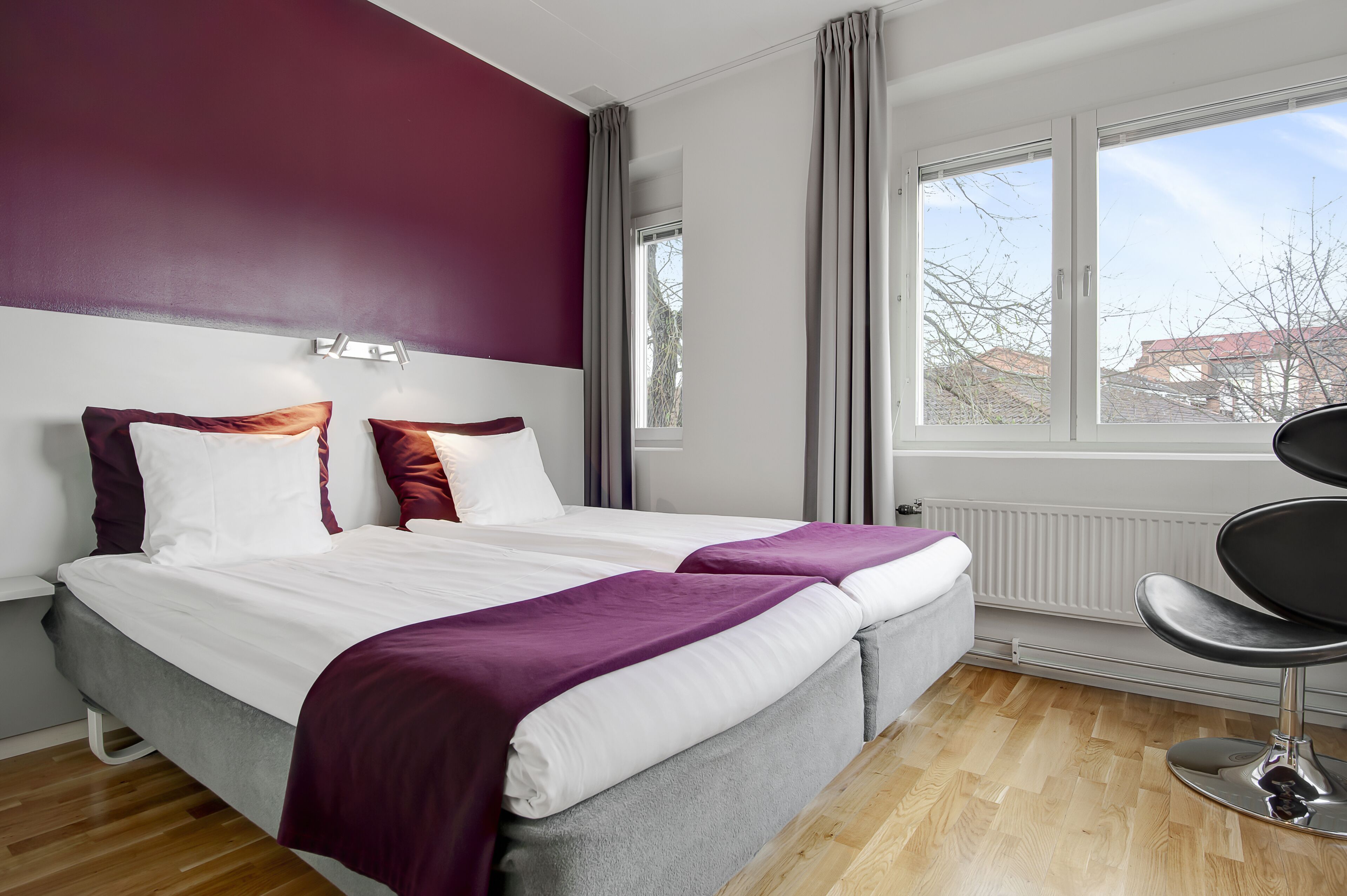 Une chambre d’hôtel moderne avec un grand lit, deux tables de chevet, une chaise et une fenêtre donnant sur des immeubles.