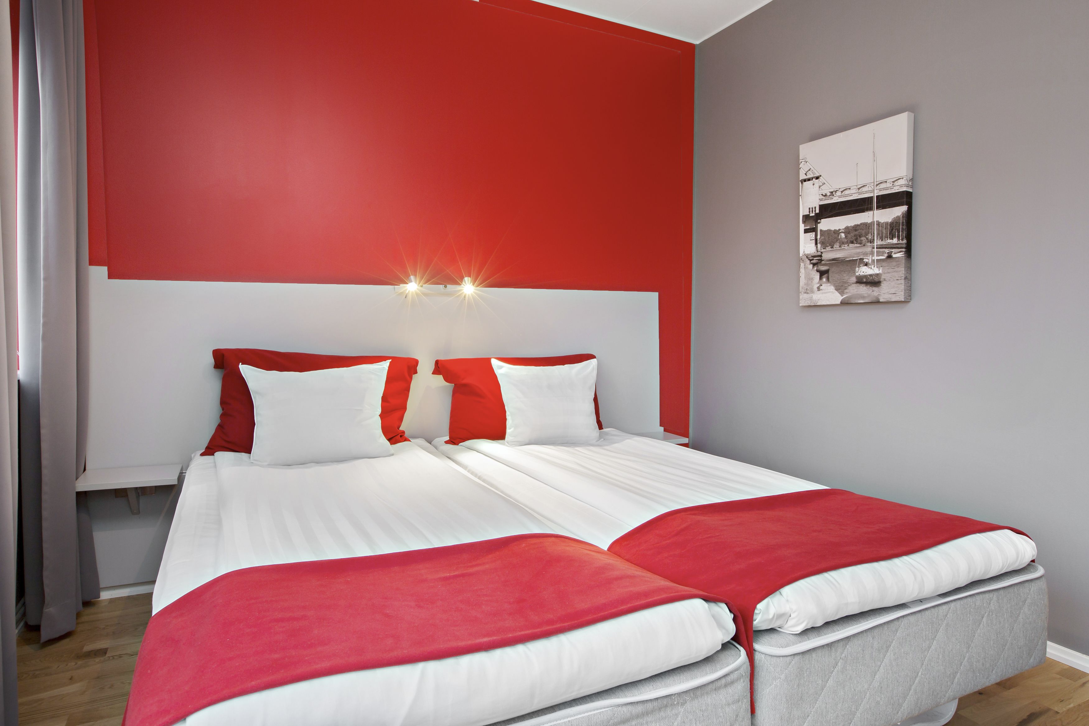 A modern hotel room with a large bed, red and white bedding, a headboard with lights, and a framed picture on the wall.