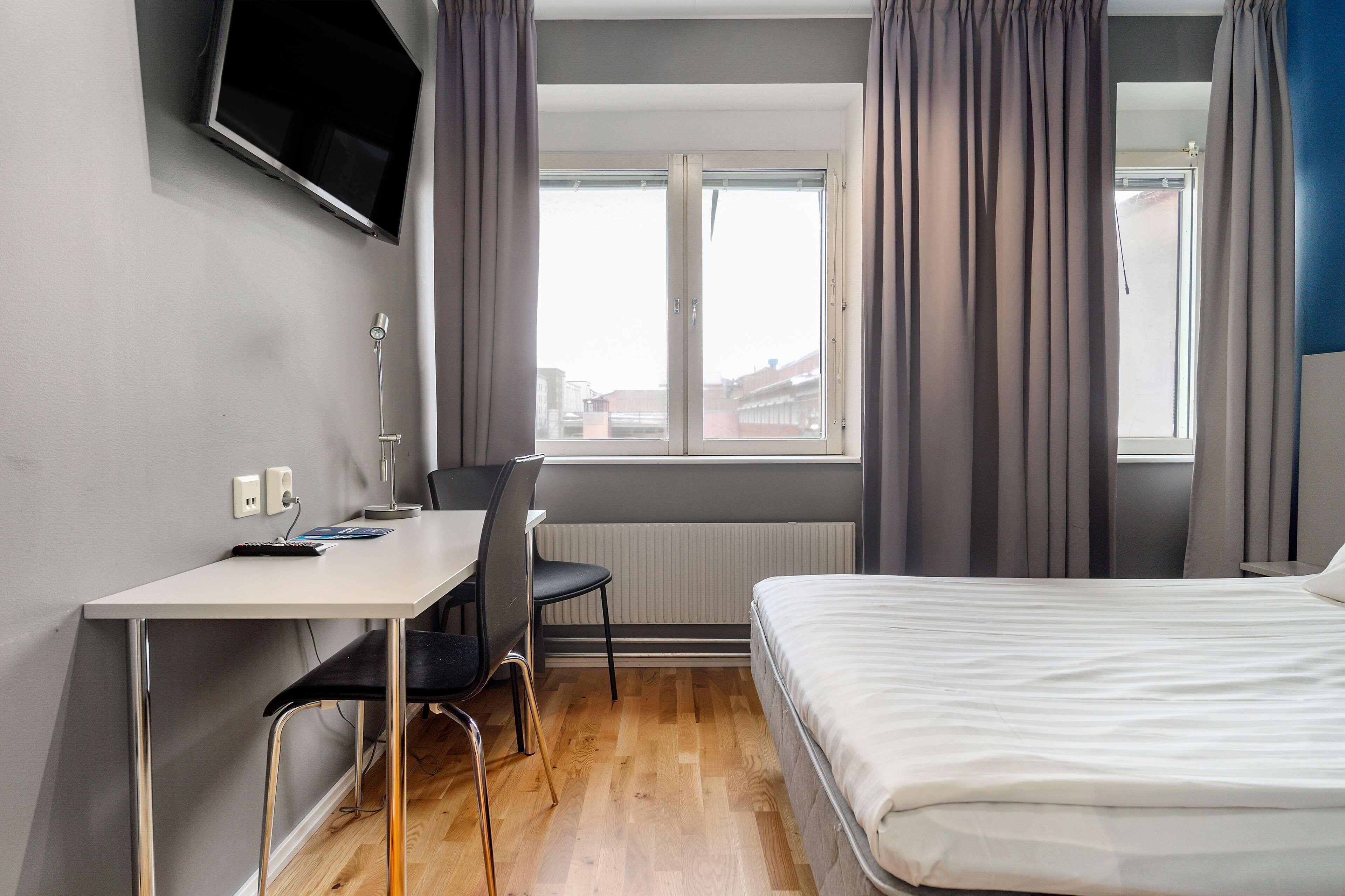 A bedroom with a bed, a desk, and a chair. There is a window with curtains and a view of buildings outside.