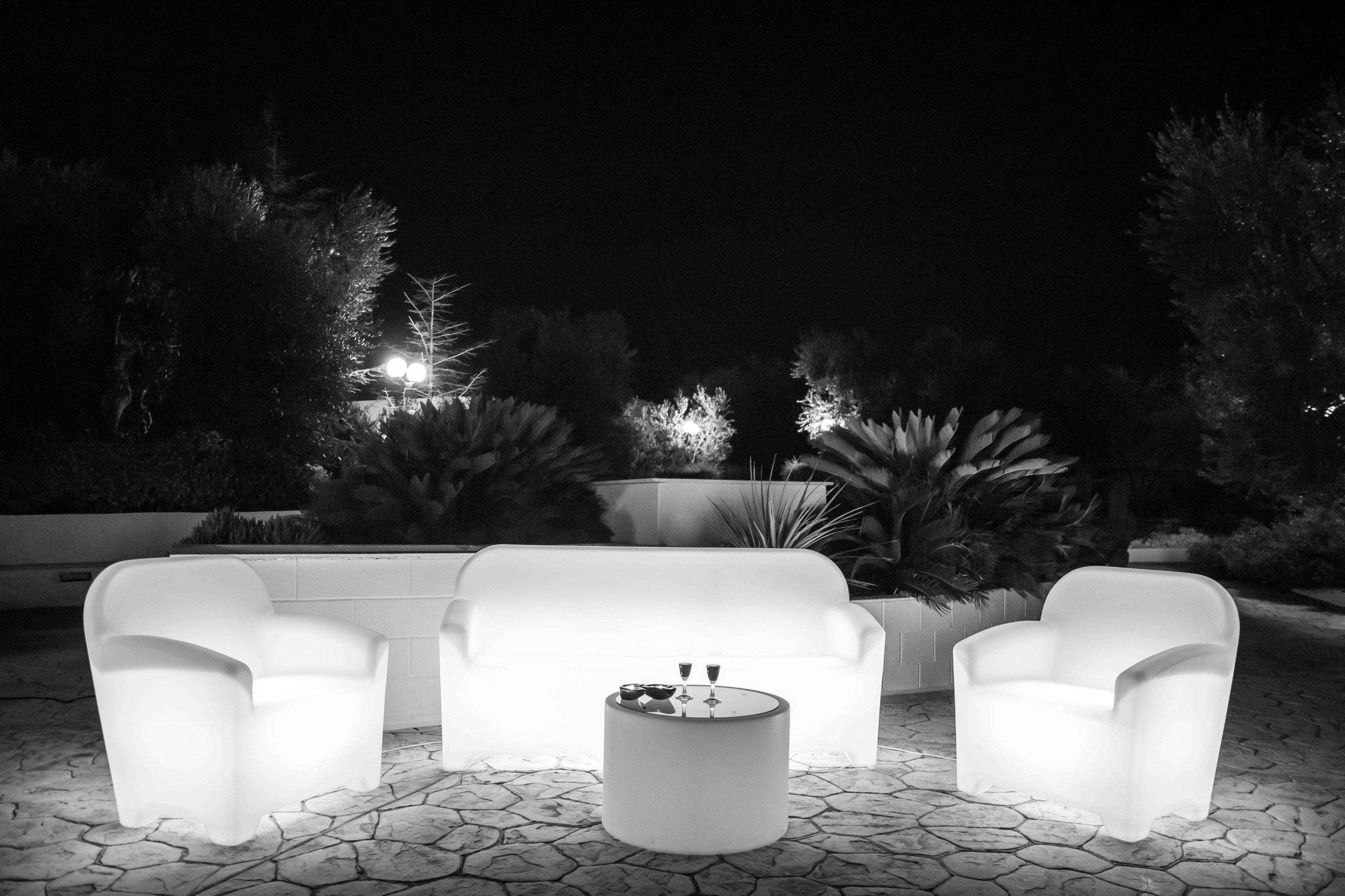 An outdoor seating area with white chairs and a round table, surrounded by potted plants and a white wall.
