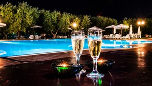Une table placée au bord de la piscine, avec deux coupes à champagne et une bouteille, entourée de chaises longues et de parasols.