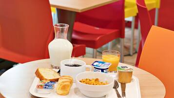 A breakfast tray with toast, yogurt, cereal, and juice on a table in a brightly colored dining area.