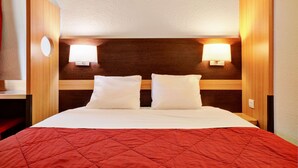 Une chambre d’hôtel avec une tête de lit en bois, deux lampes de chevet, une couvre-lit rouge matelassée et un oreiller blanc.