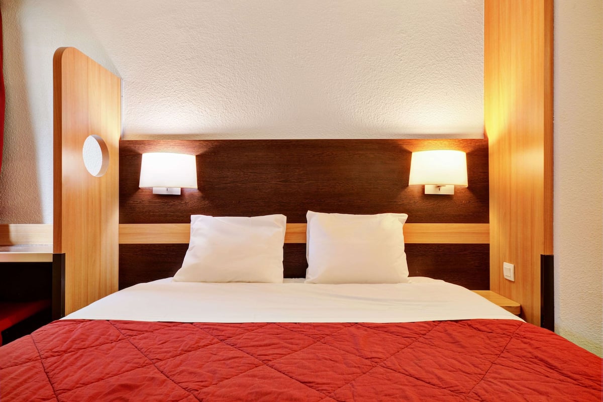 A hotel room with a wooden headboard, two bedside lamps, a red quilted bedspread, and a white pillow.