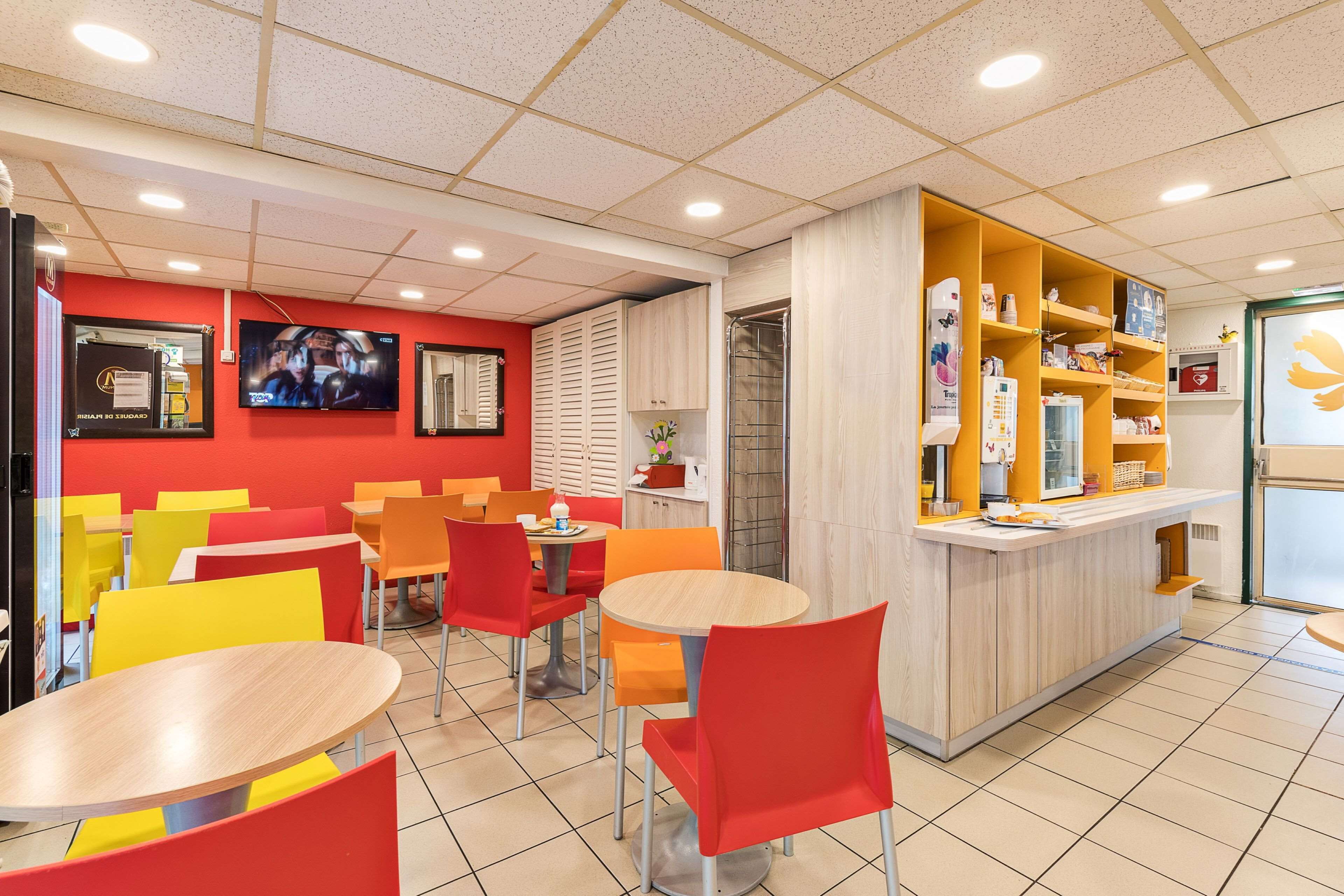 Un restaurante bien iluminado con sillas rojas y amarillas, una barra de madera y un televisor en la pared.