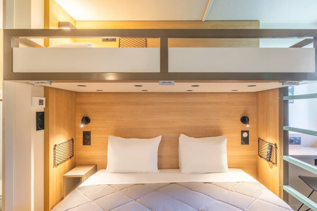 Una cama litera con cabecera de madera, dos almohadas, mesitas de noche con parlantes y una ventana con vistas.