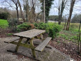 Une table de pique-nique en bois, située dans un parc, avec des arbres et des arbustes en arrière-plan.