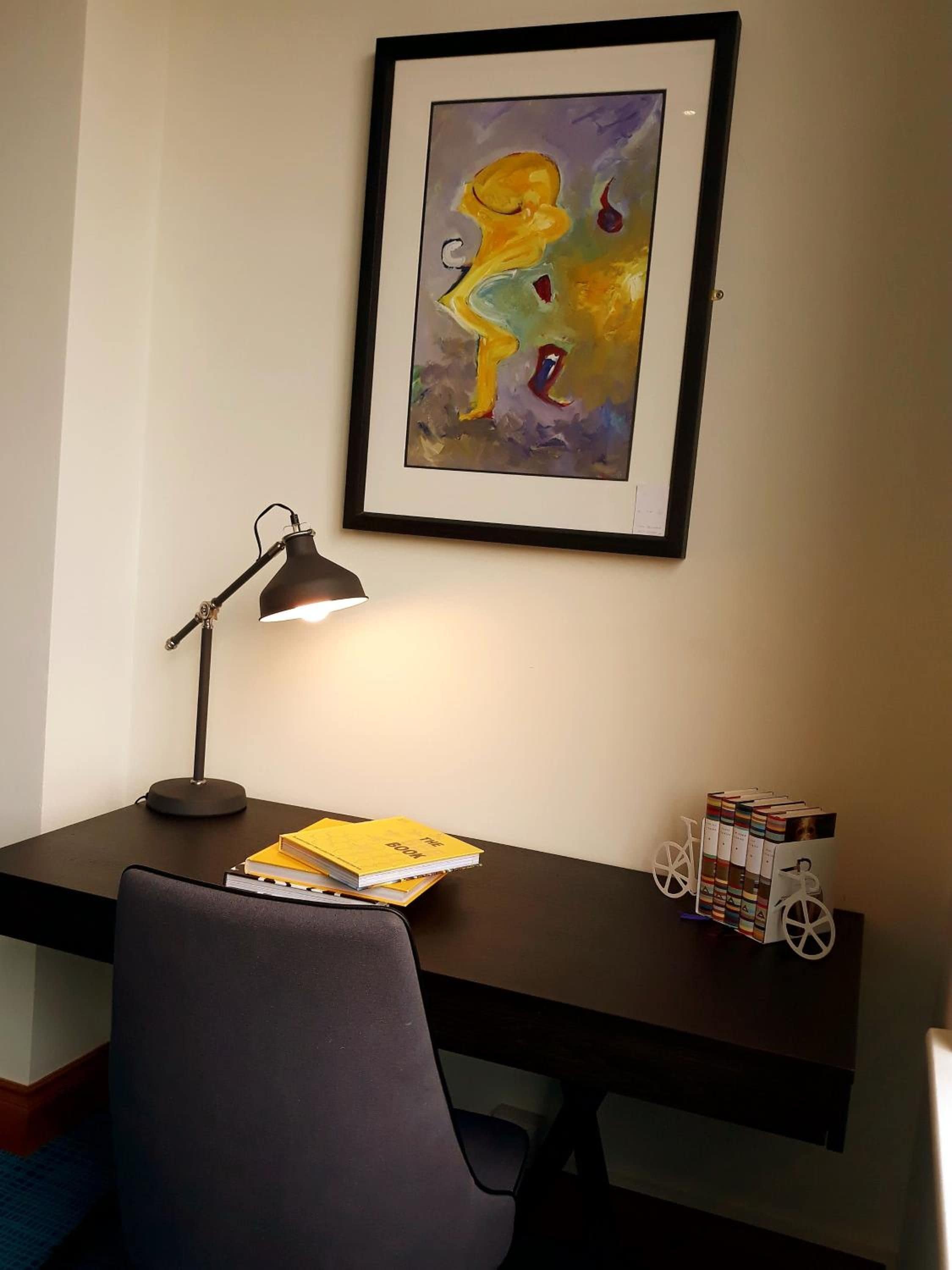 Un bureau avec une lampe, une chaise, un tableau abstrait encadré et une bibliothèque remplie de livres.