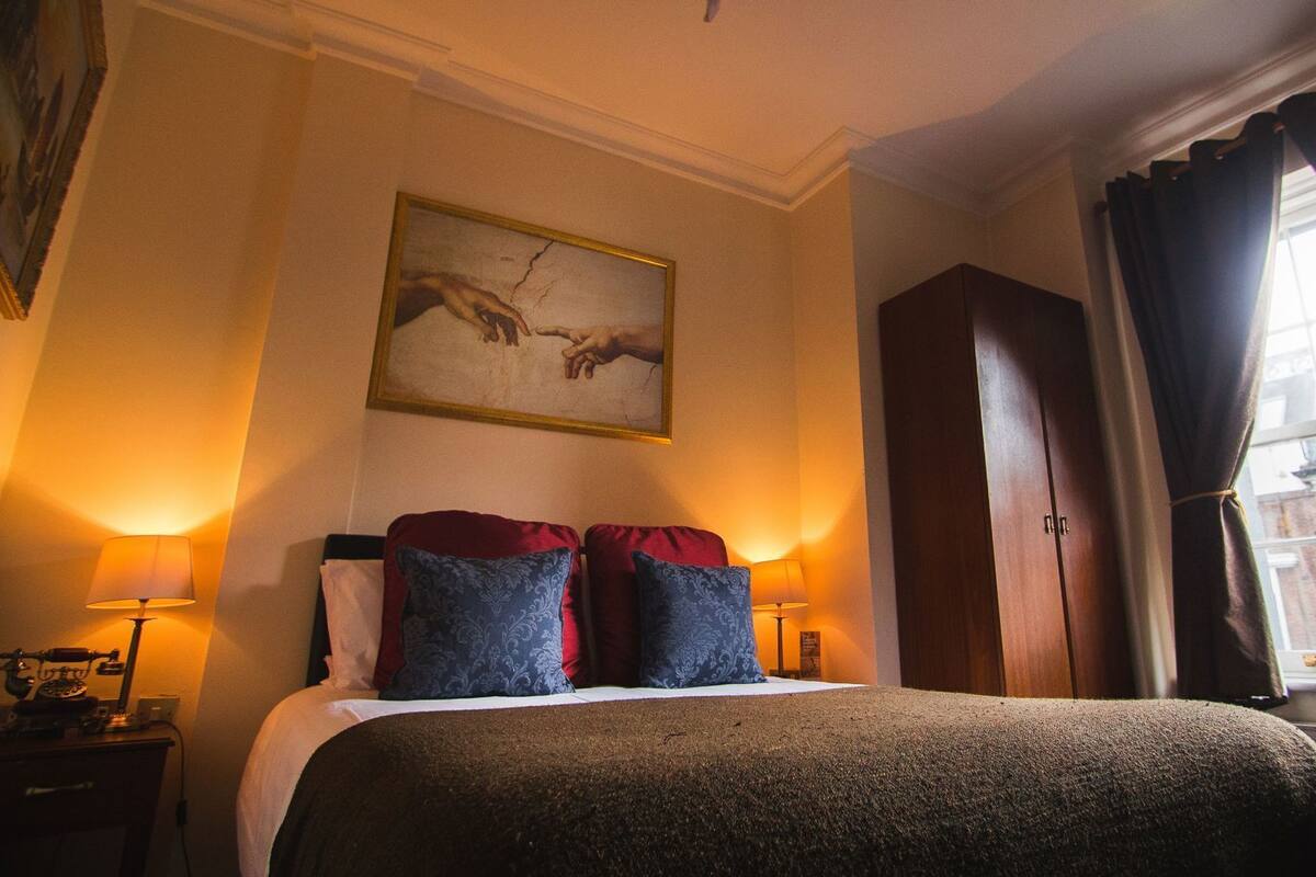 A hotel room with a bed, two lamps, a painting, a window with curtains, and a view of a building.