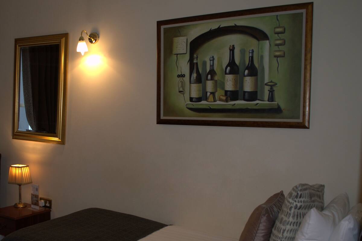 A hotel room with a bed, a nightstand with a lamp, a framed painting of wine bottles, and a mirror.
