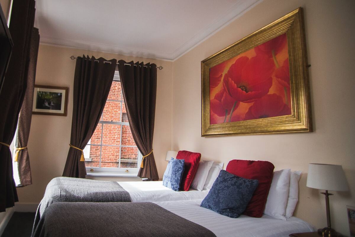A hotel room with two beds, a window with curtains, a painting on the wall, and a lamp on the nightstand.