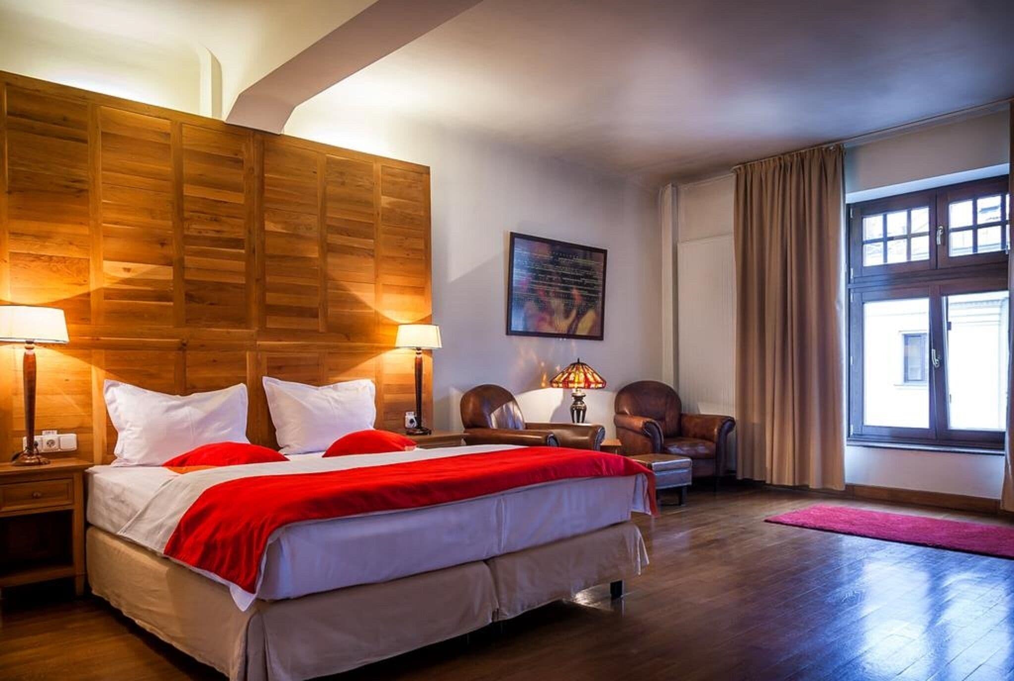 A hotel room with a large bed, wooden headboard, two bedside lamps, a desk with a chair, a window with curtains, and a wall-mounted artwork.