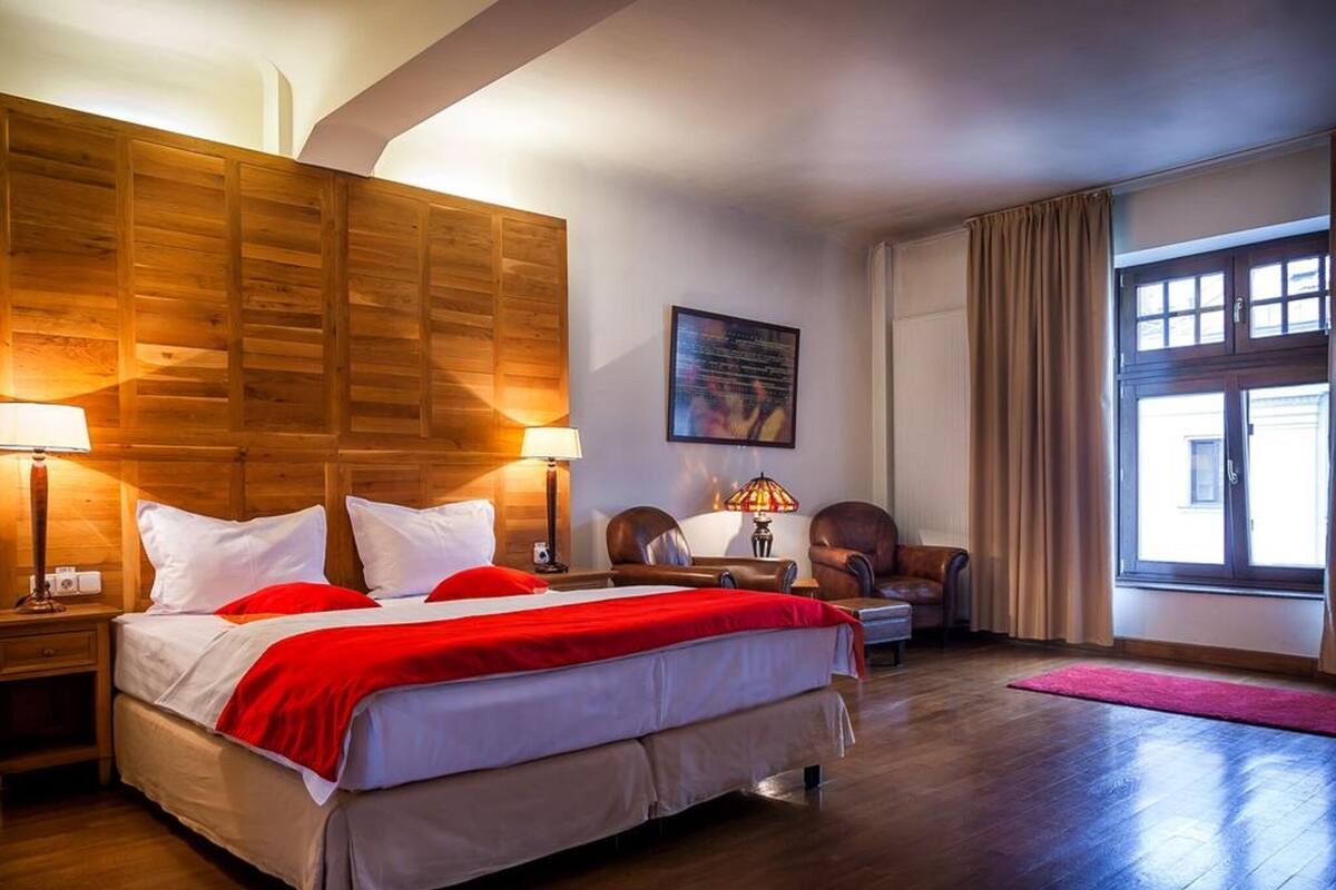 A hotel room with a large bed, wooden headboard, two bedside lamps, a desk with a chair, a window with curtains, and a wall-mounted artwork.