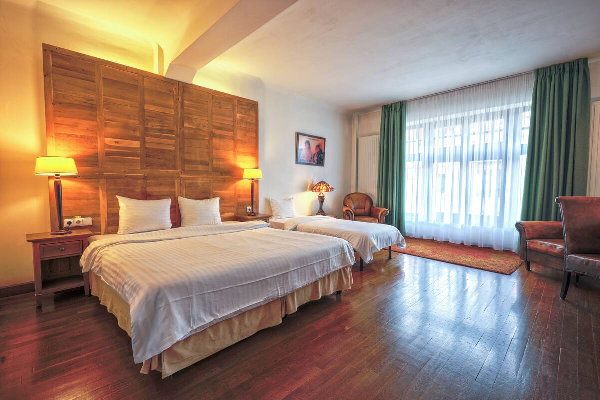 A hotel room with two beds, wooden headboard, two bedside lamps, a chair, and a large window with curtains.