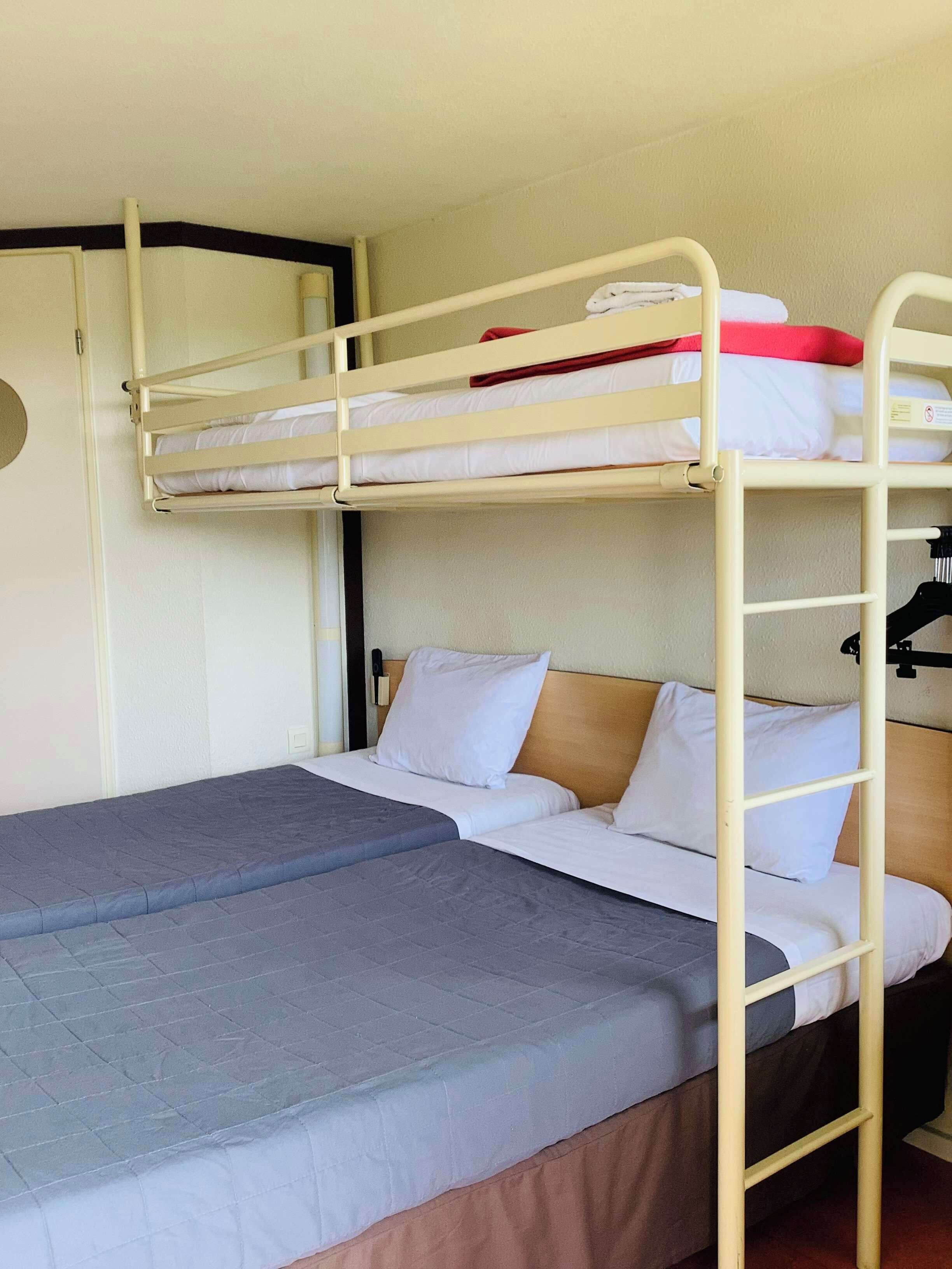 Ein Schlafraum mit Etagenbett, einem unteren Bett und einem Einzelbett. Eine Leiter ist am unteren Bett befestigt, und rechts ist ein kleiner Schreibtisch mit Stuhl zu sehen.