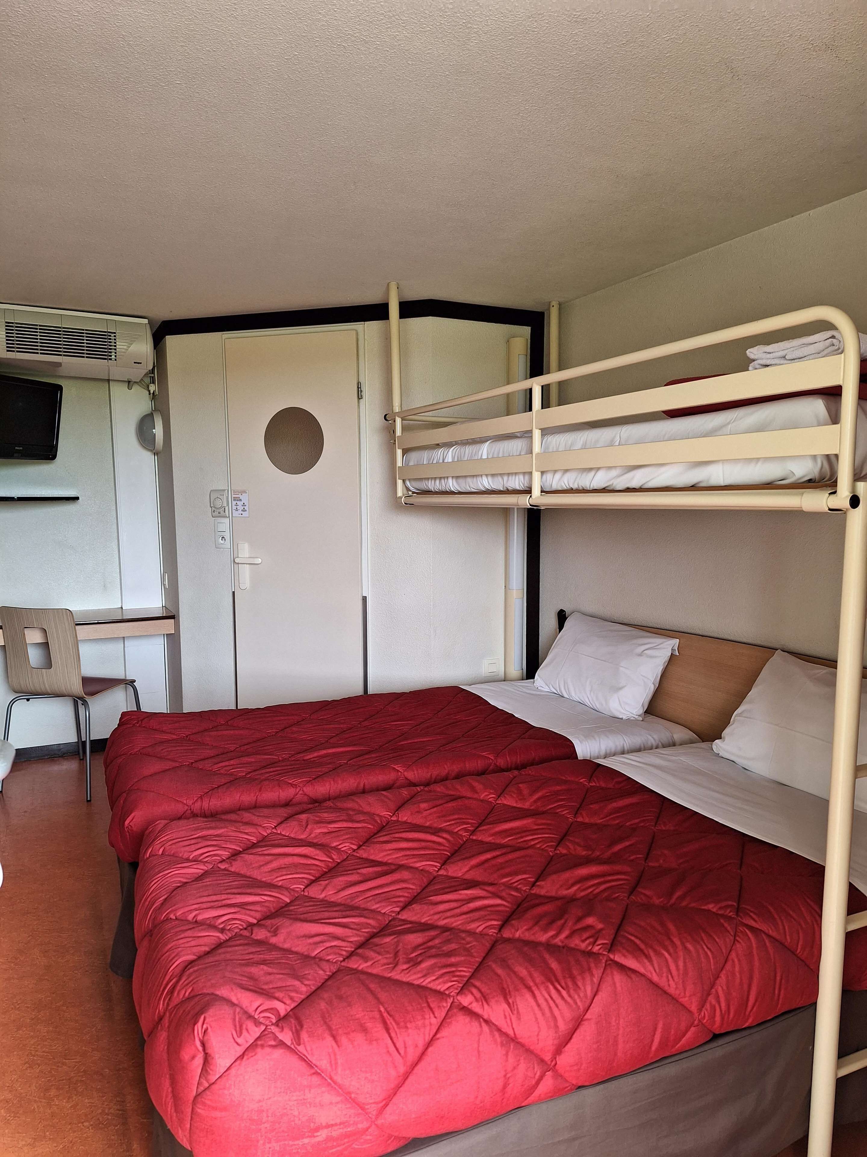 A bunk bed with a red quilt, a desk with a chair, and a TV mounted on the wall.