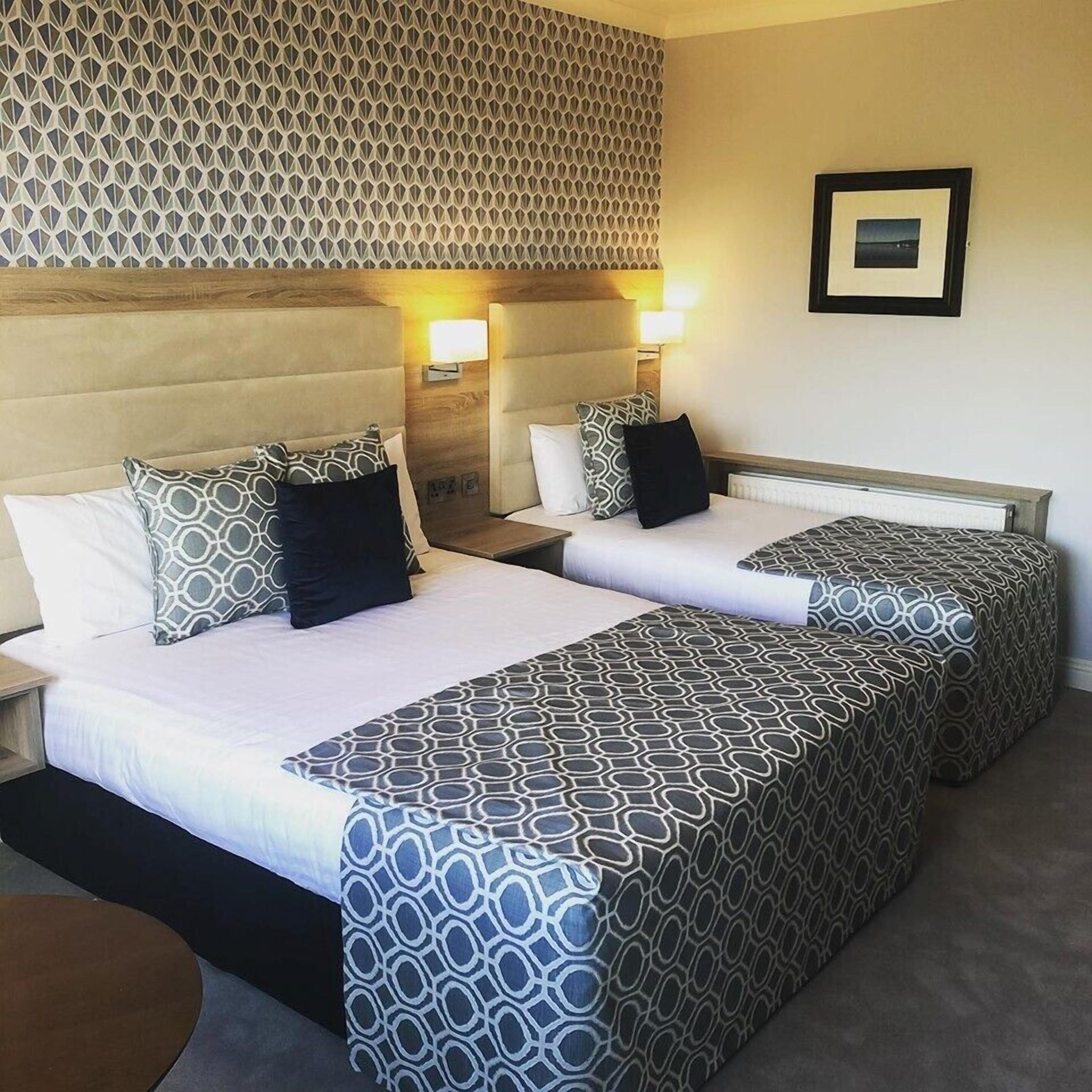 A hotel room with two beds, each with patterned bedding and pillows, a wooden headboard, and a wall-mounted picture.