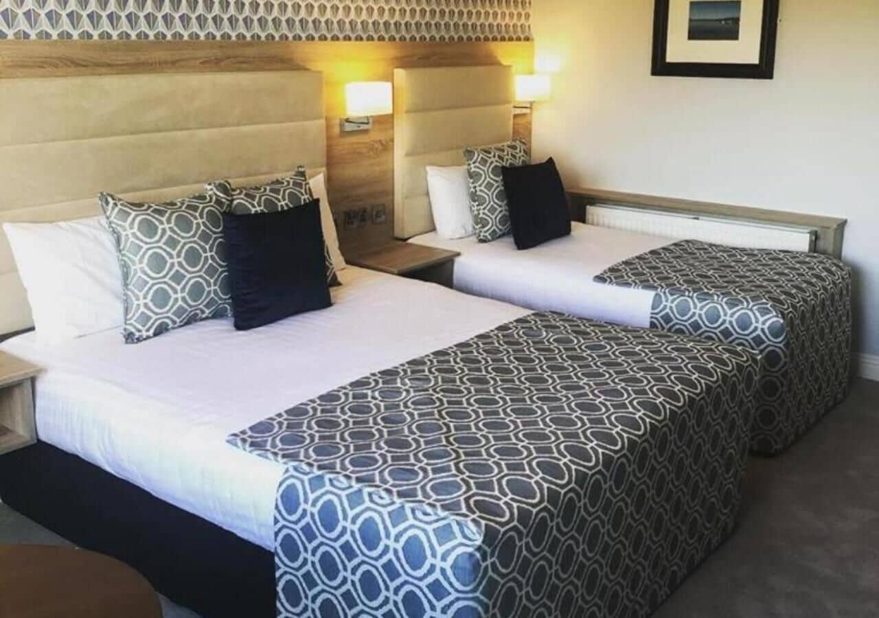 A hotel room with two beds, each with patterned bedding and pillows, a wooden headboard, bedside lamps, and a framed picture on the wall.
