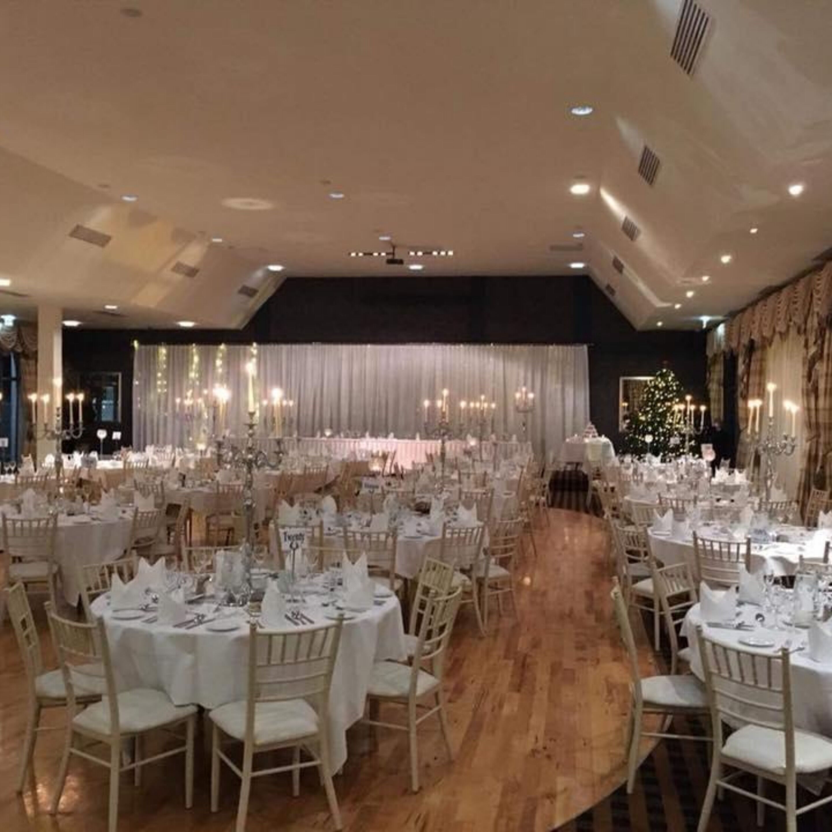 A banquet hall with round tables set for events, a stage with draped curtains, and a Christmas tree.