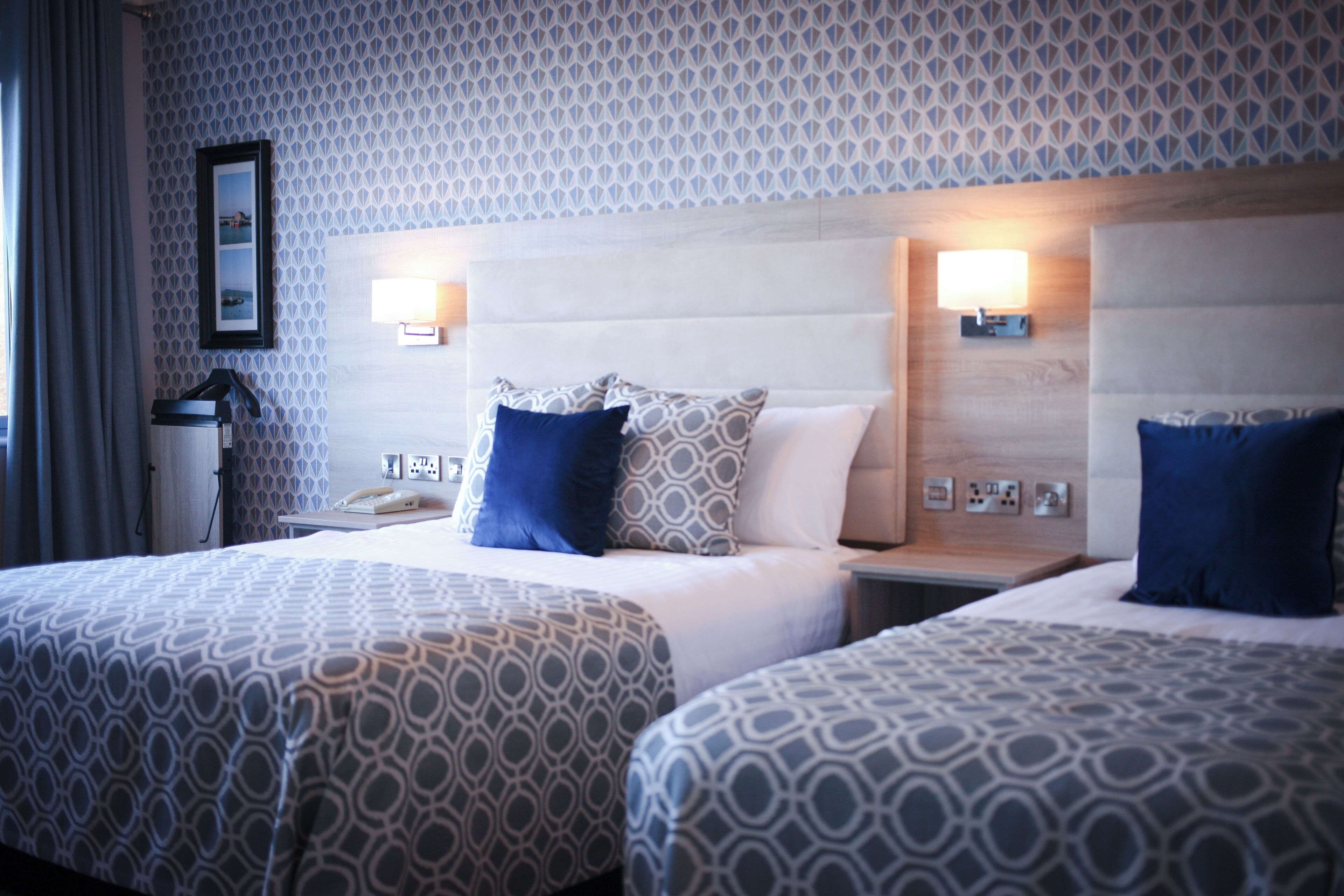 A hotel room with two beds, patterned bedding, bedside lamps, and a wall-mounted phone.