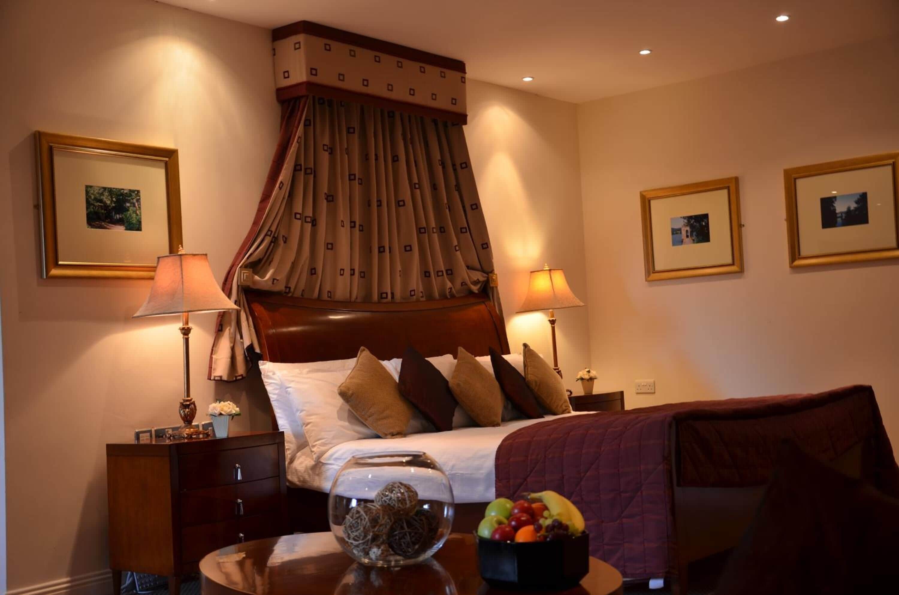 A hotel room with a large bed, two bedside tables, a nightstand, a decorative lamp, a vase with flowers, a fruit bowl, and a framed picture on the wall.