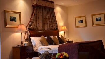 A hotel room with a large bed, two bedside tables, a nightstand, a decorative lamp, a vase with flowers, a fruit bowl, and a framed picture on the wall.