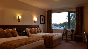 A hotel room with two beds, a small table, and a chair. There is a window with curtains and a view of trees and buildings outside.