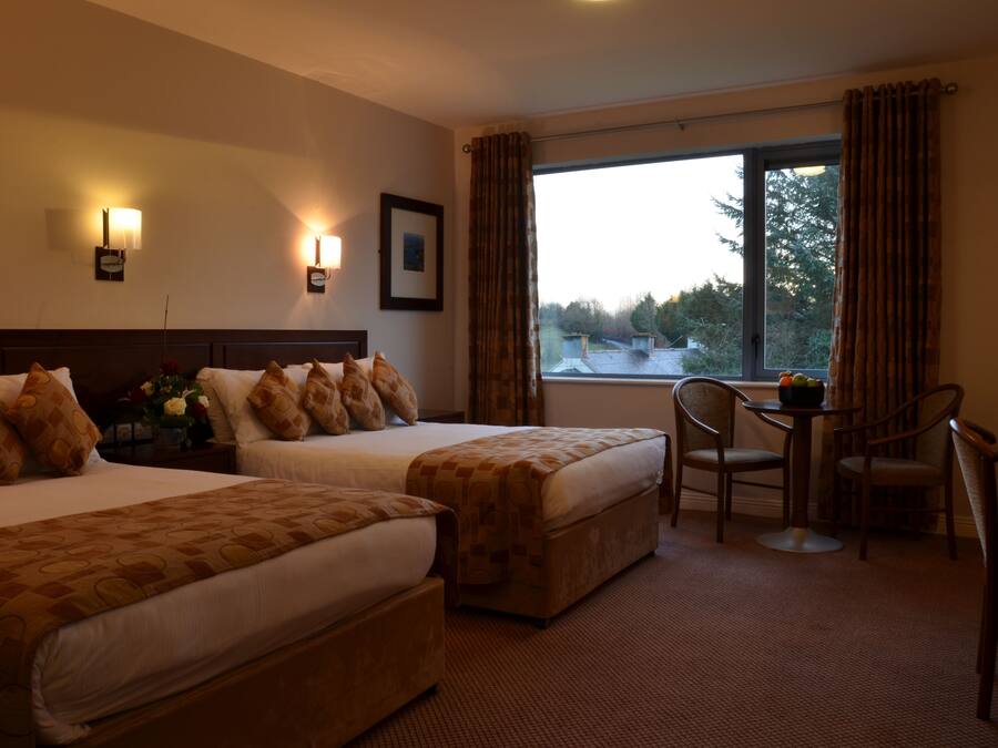 A hotel room with two beds, a small table, and a chair. There is a window with curtains and a view of trees and buildings outside.