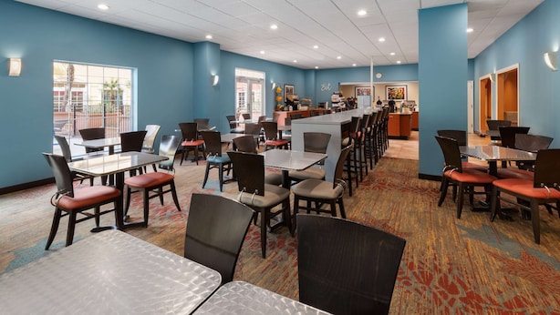 A hotel dining area with multiple tables and chairs, a counter with a coffee machine, and a view of the outside through large windows.
