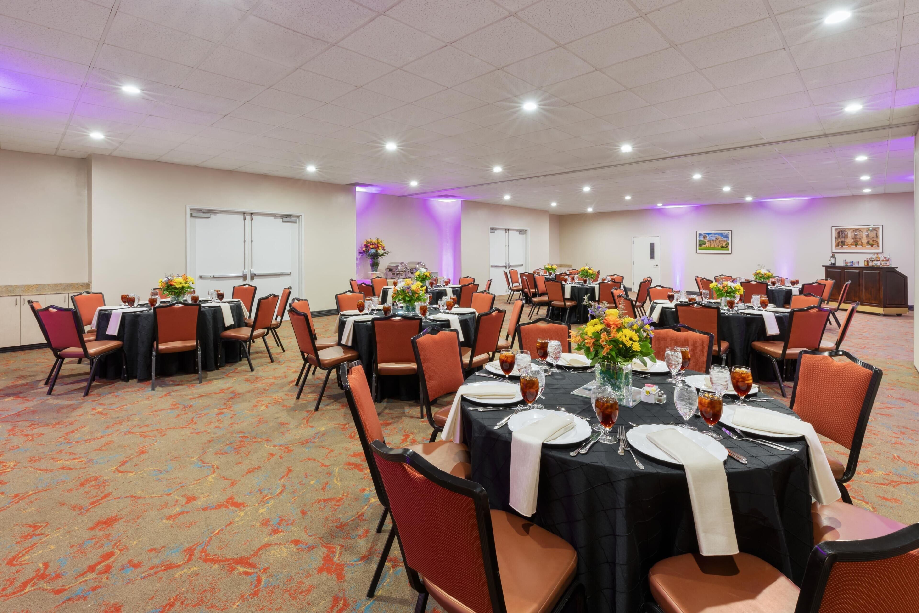 Une salle de conférence avec plusieurs tables dressées pour un repas, équipée de chaises, de tables et de centres de table.