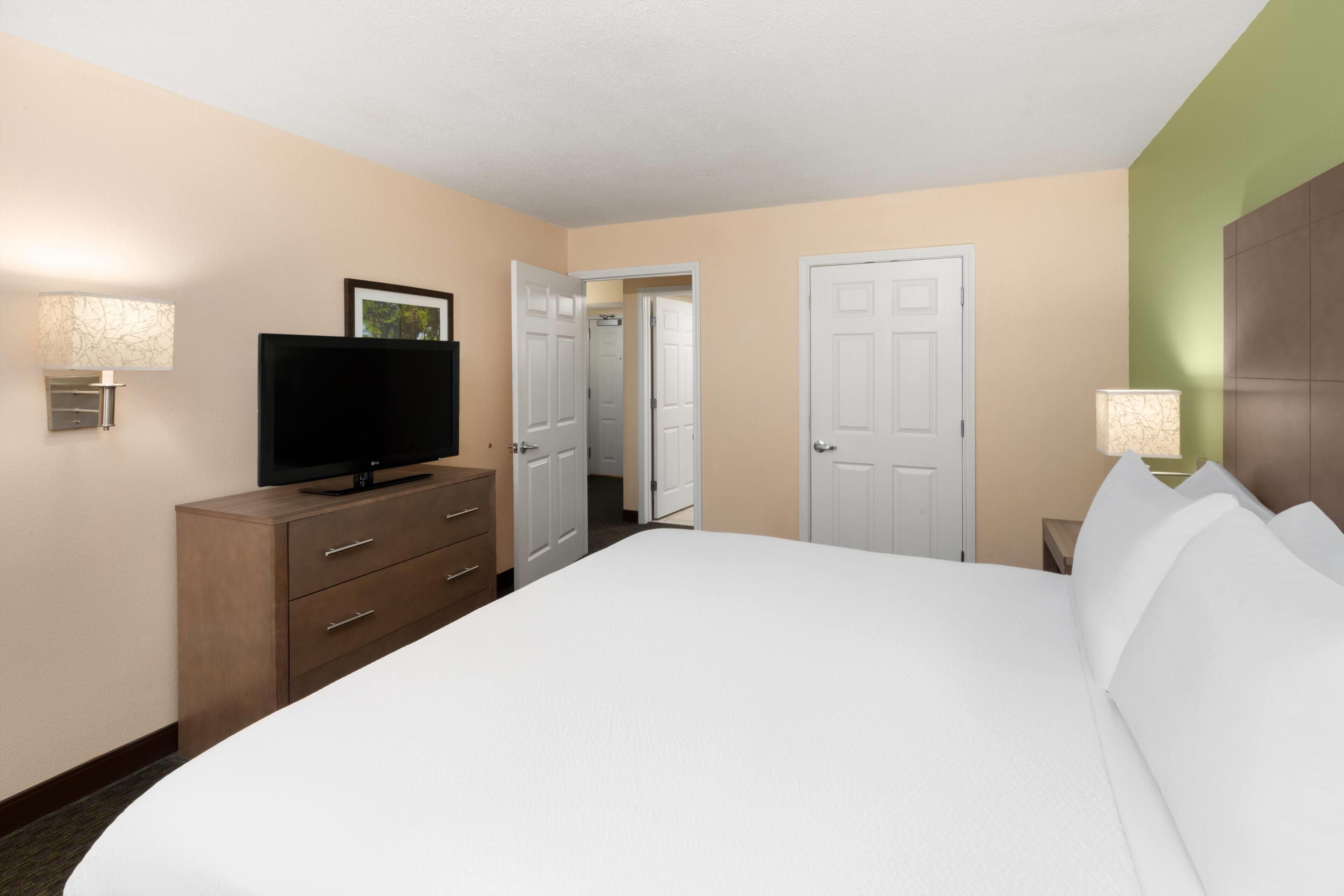 A hotel room with a bed, a television on a wooden dresser, a door, and a lamp.