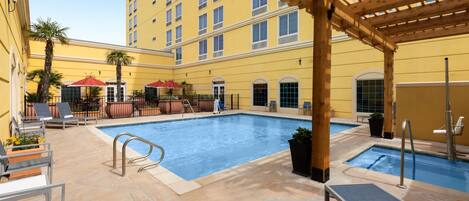 A hotel pool area with lounge chairs, a pergola, and a building with multiple windows.